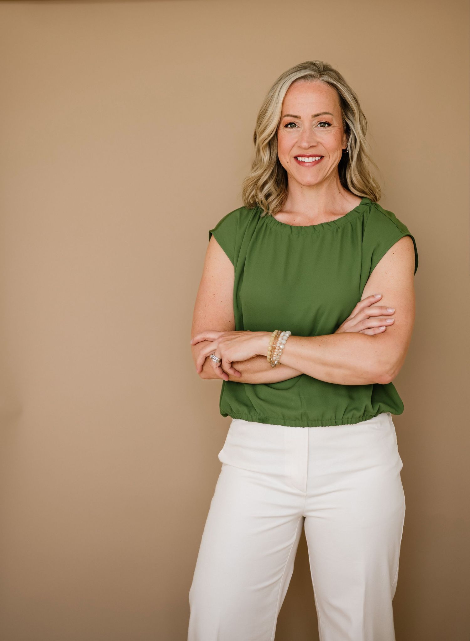 Professional portrait of someone in a green sleeveless blouse and white pants standing confidently against beige background.