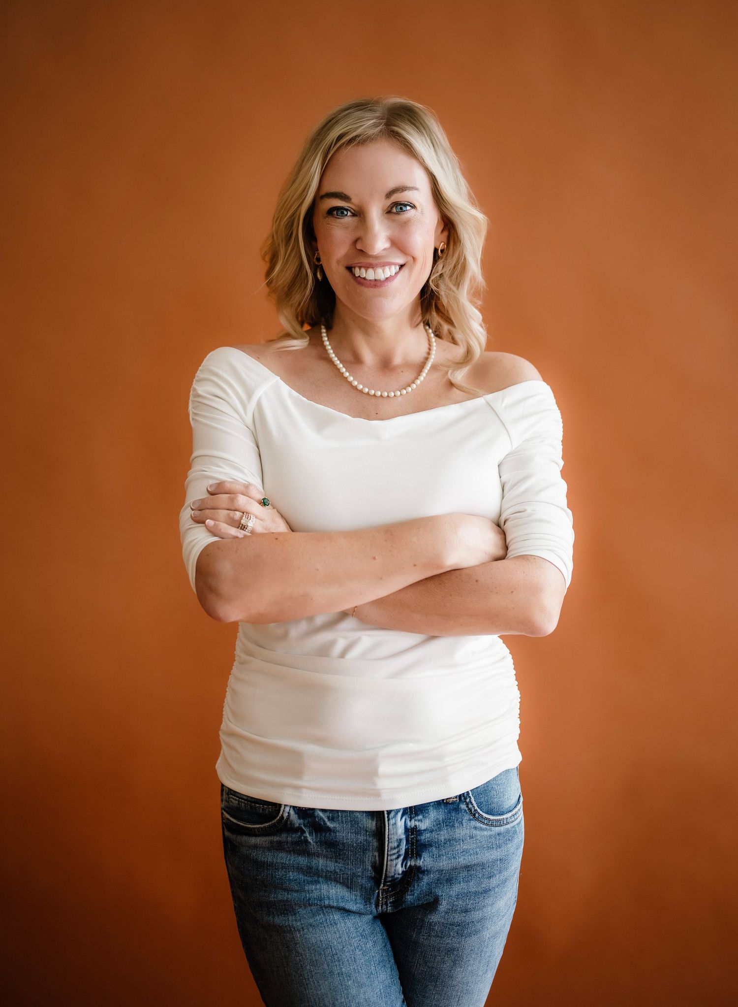 A person in a white long-sleeve top and blue jeans stands with arms crossed against an orange backdrop.