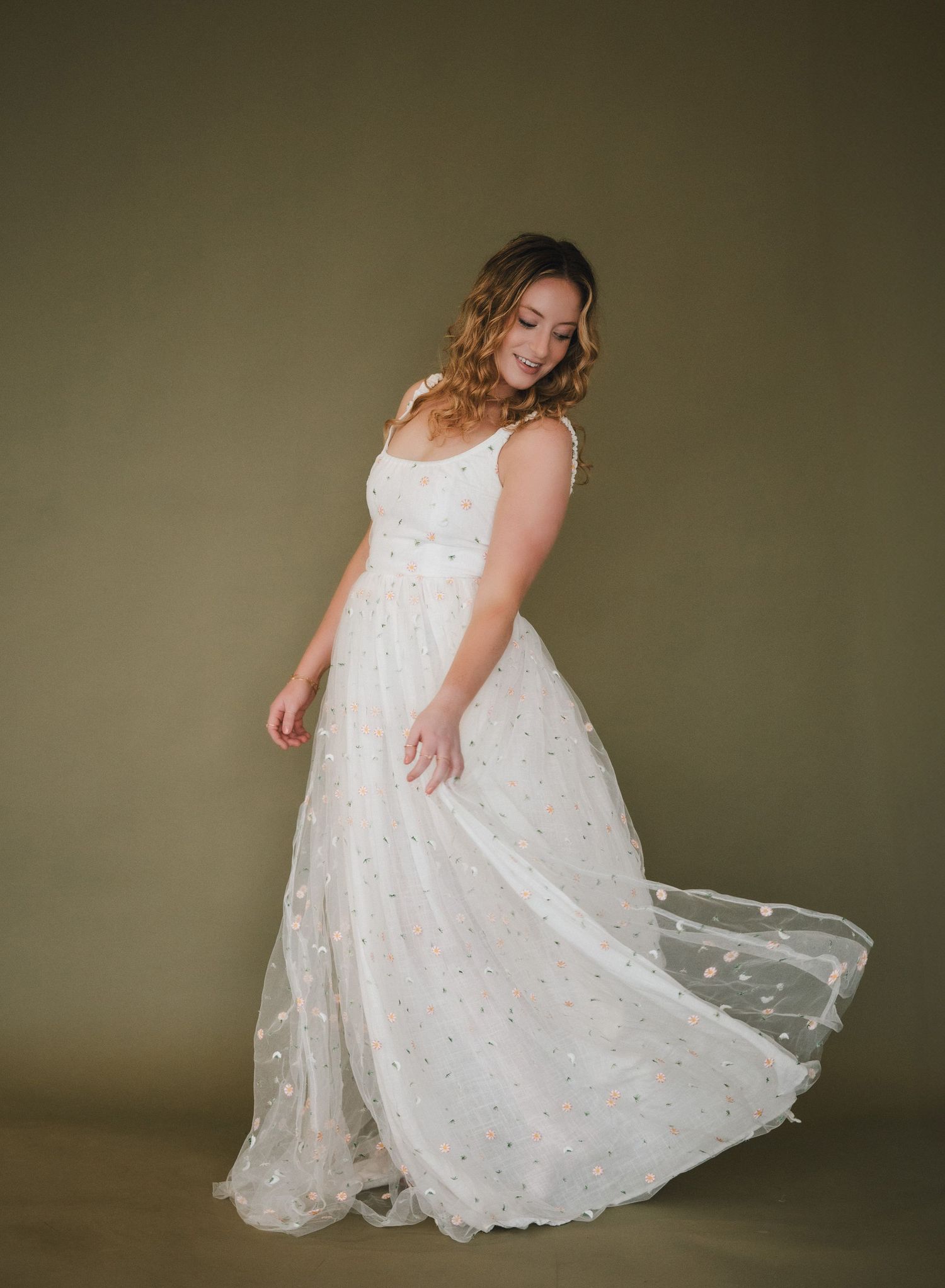 Woman twirling in a flowy white floral tulle wedding gown with spaghetti straps against an olive background.