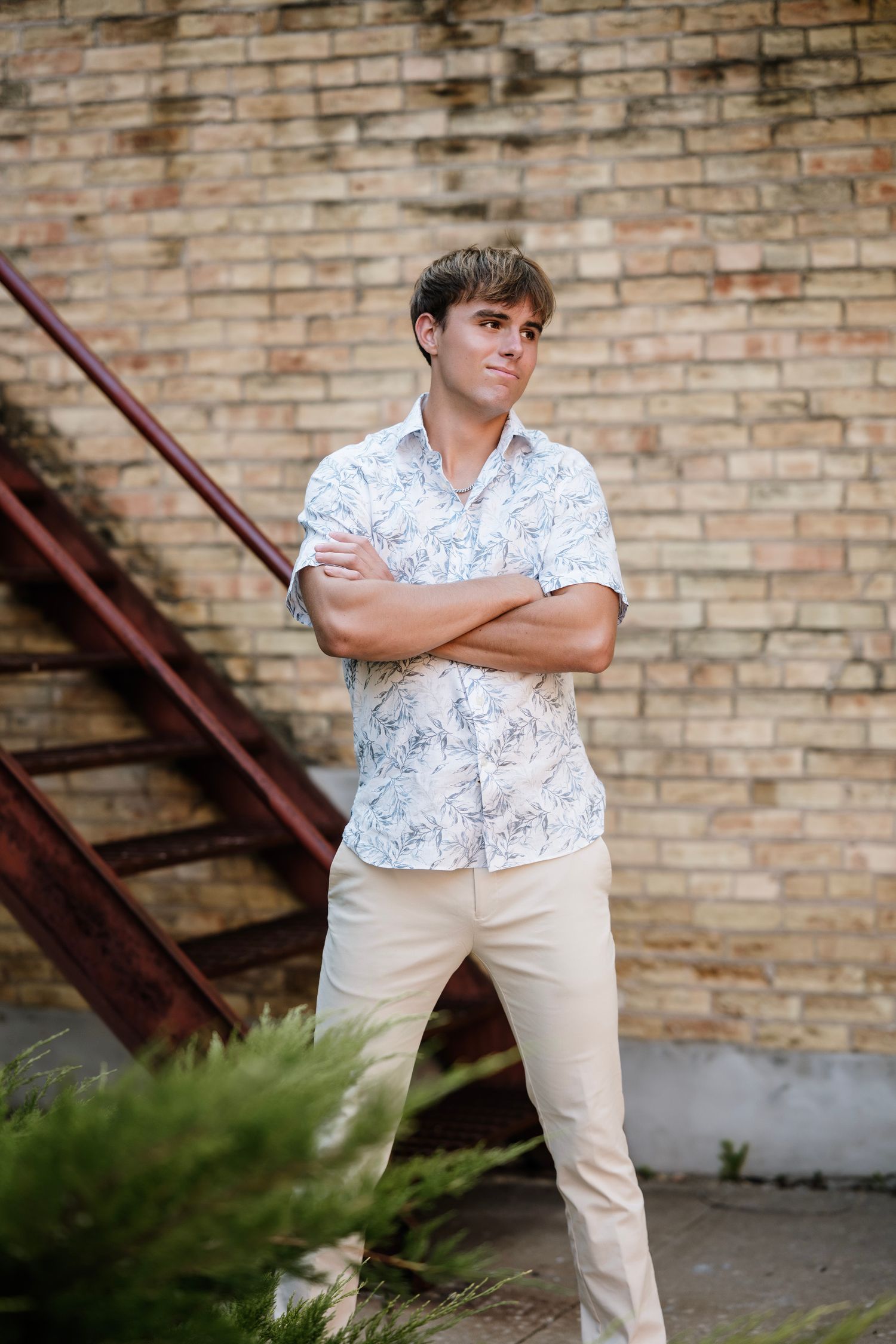 Person in white floral shirt and khaki pants poses confidently against brick wall with metal stairs.