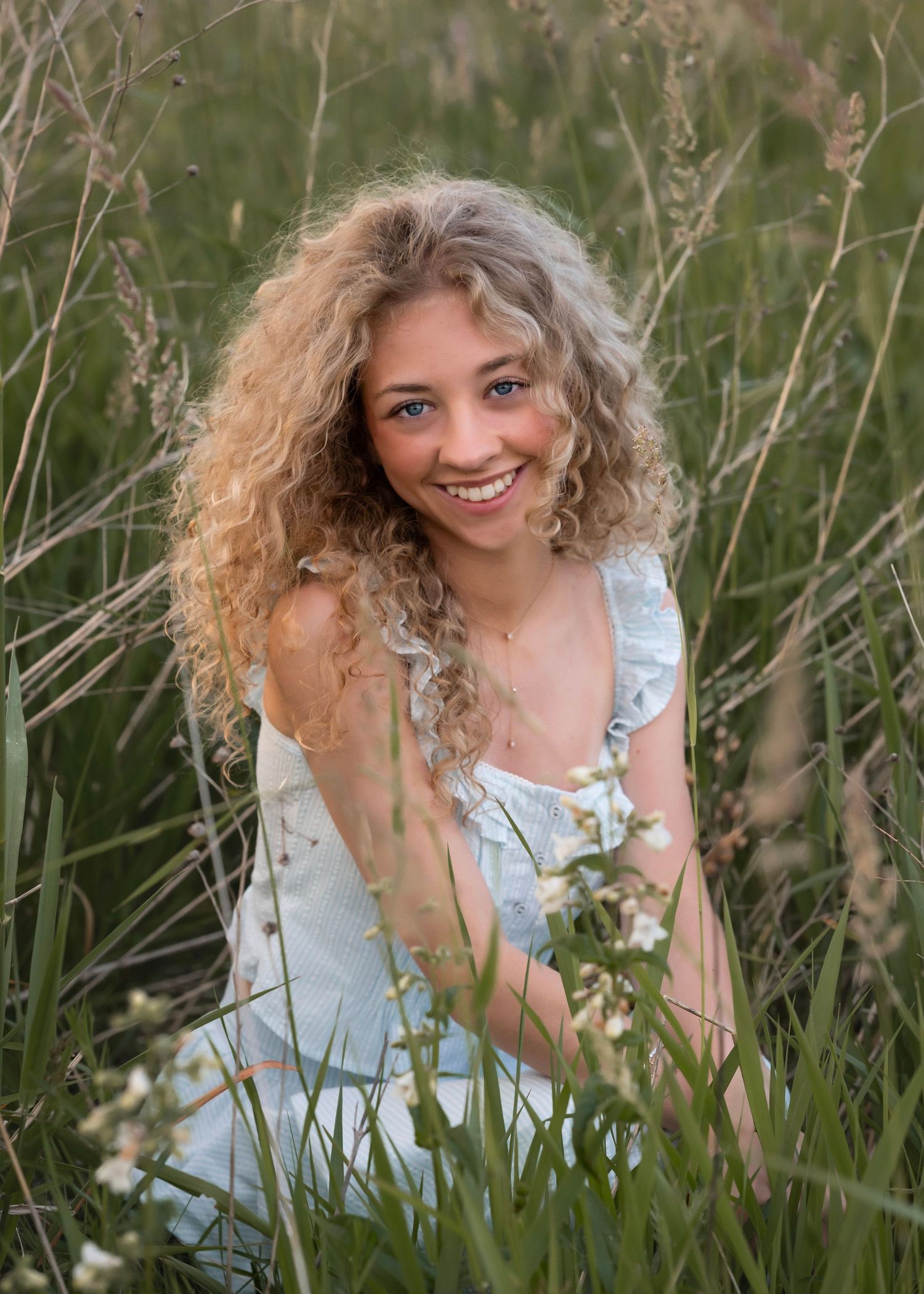 Young woman with curly blonde hair smiling in a field of tall grass and wildflowers wearing a light blue dress.