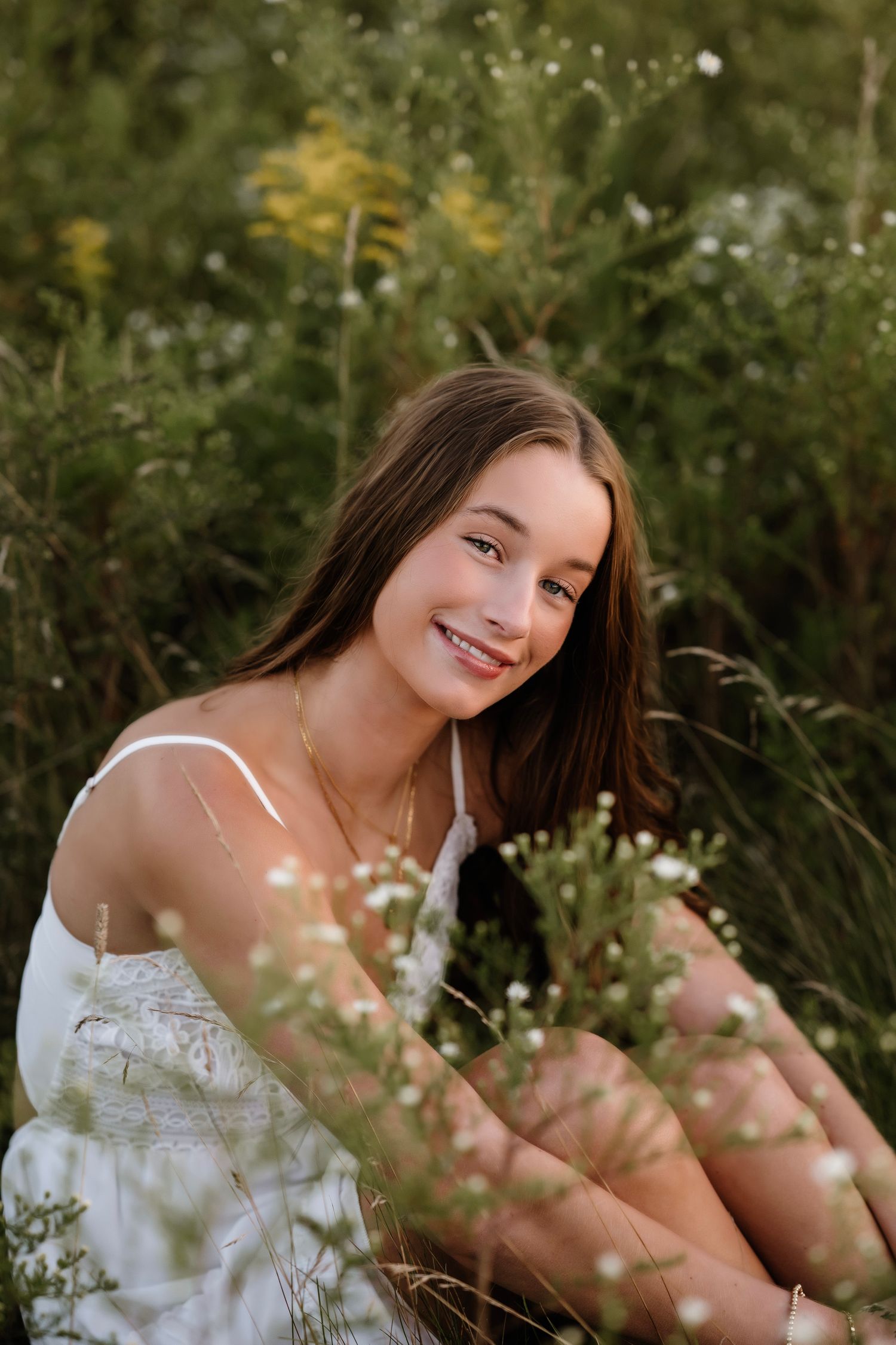 A smiling teenage girl in a white dress sits among wildflowers and green foliage in a sunny outdoor setting.