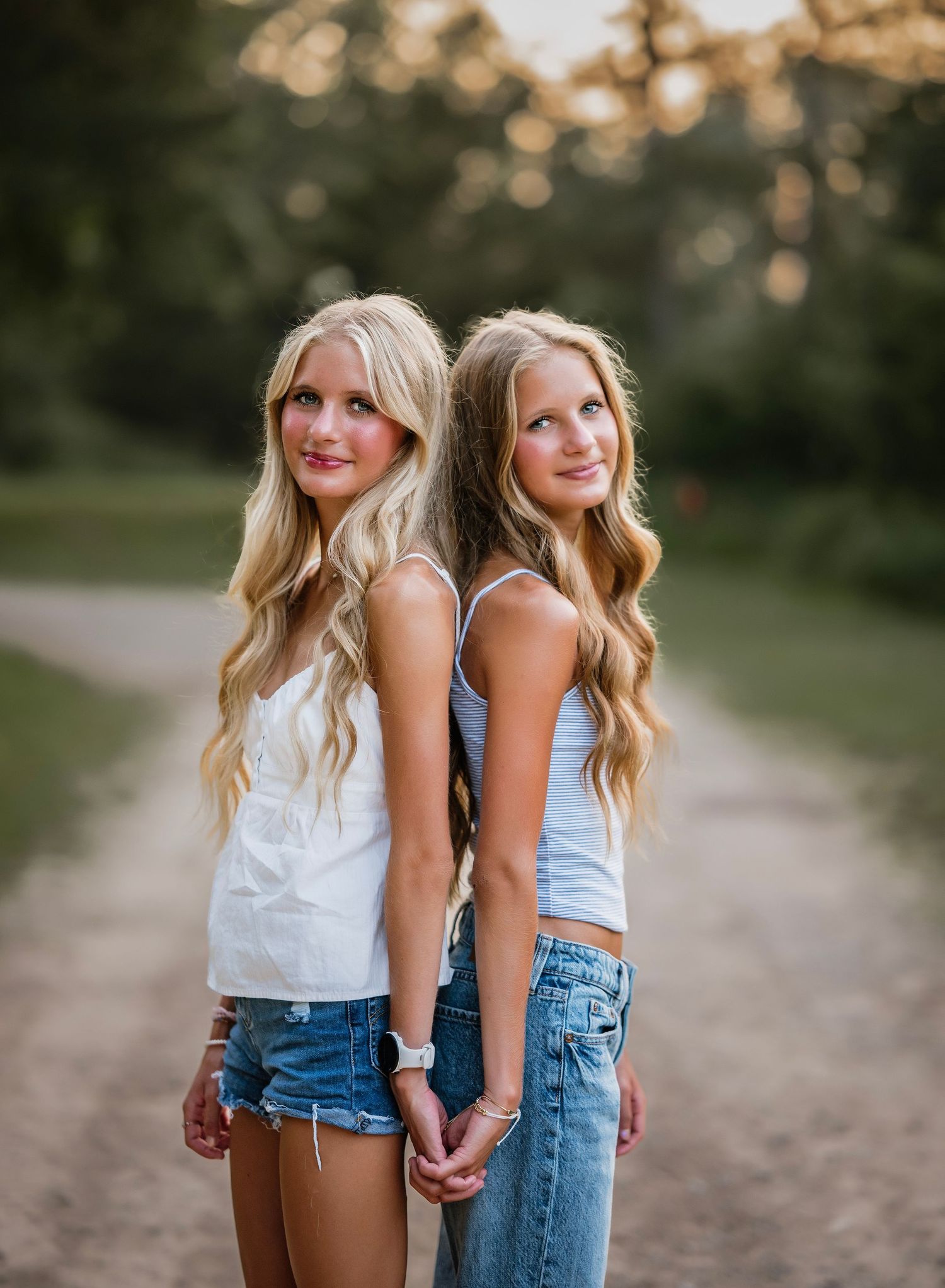 Two blonde teenage girls in denim shorts hold hands back-to-back on a scenic dirt path surrounded by lush greenery.