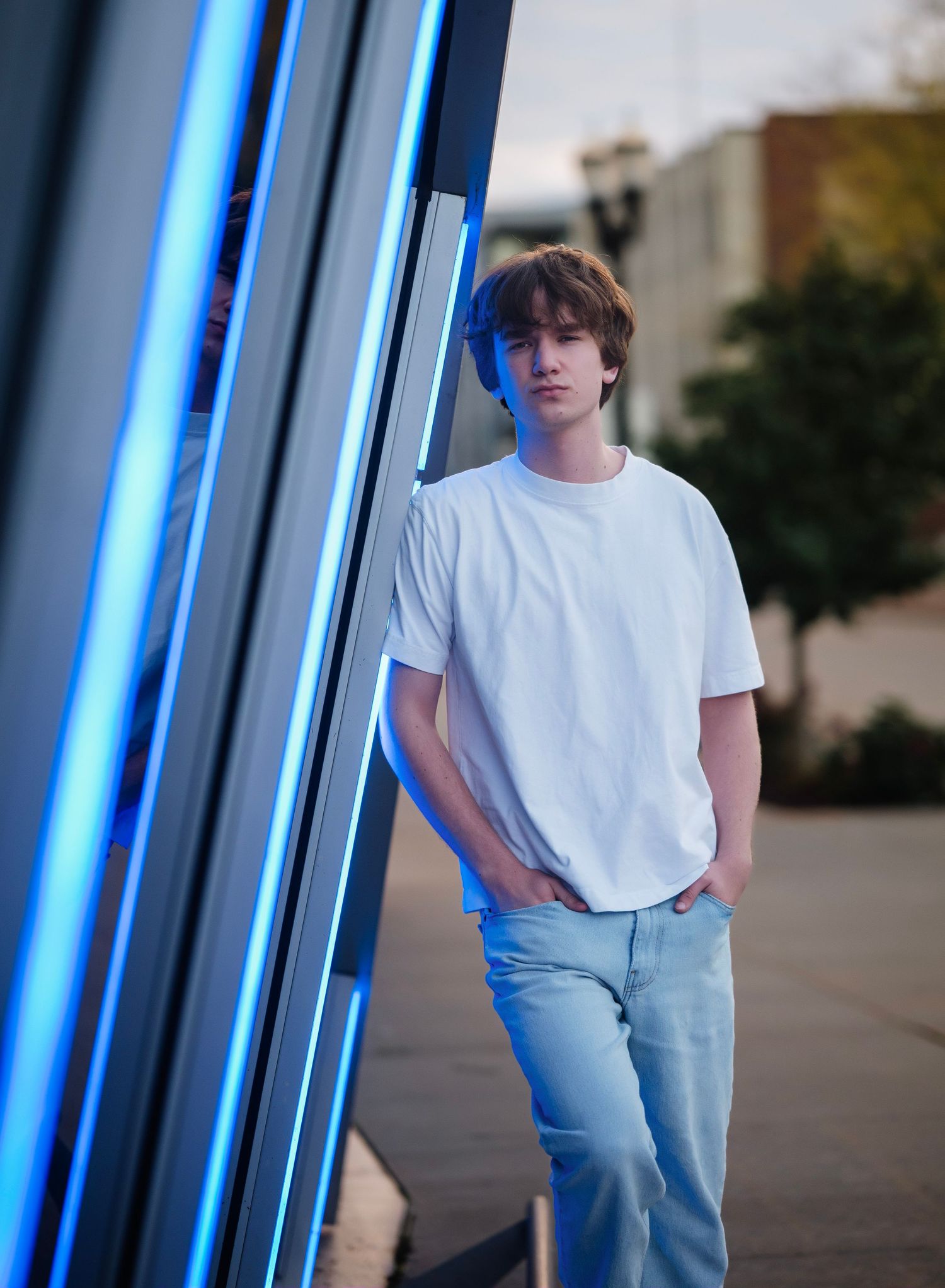Person in white t-shirt and jeans poses casually against blue neon-lit wall in urban setting.