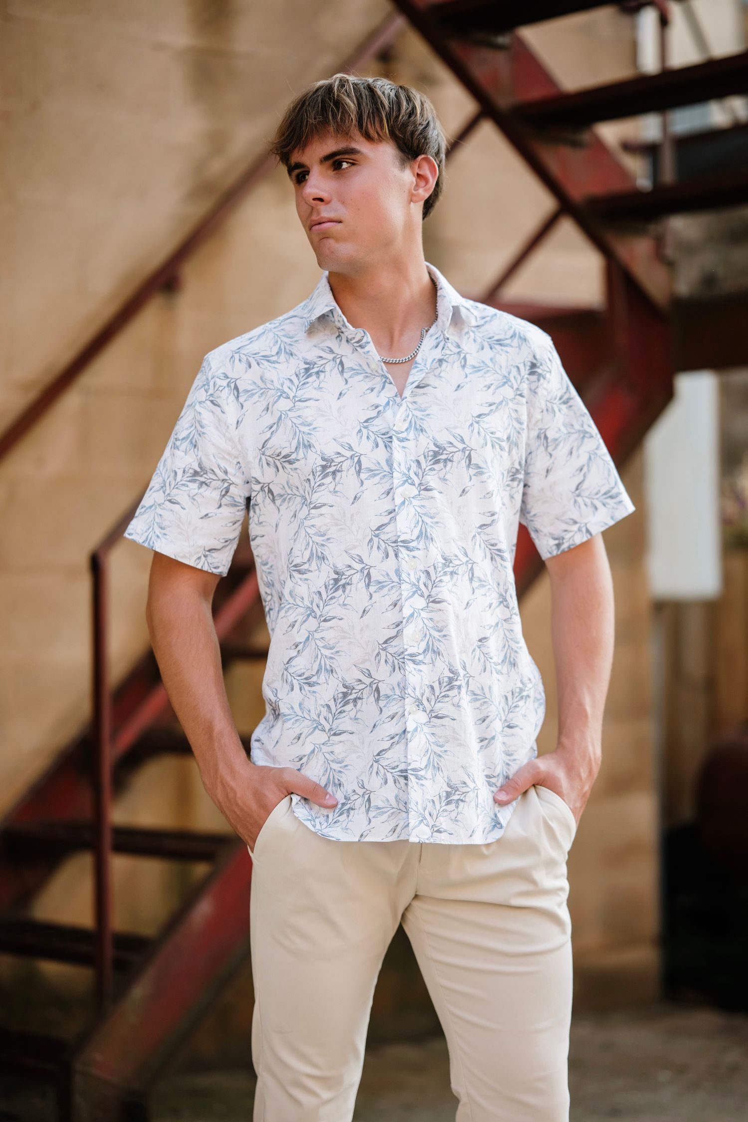 A person wearing a white floral short-sleeve button-up shirt and khaki pants stands against a staircase.