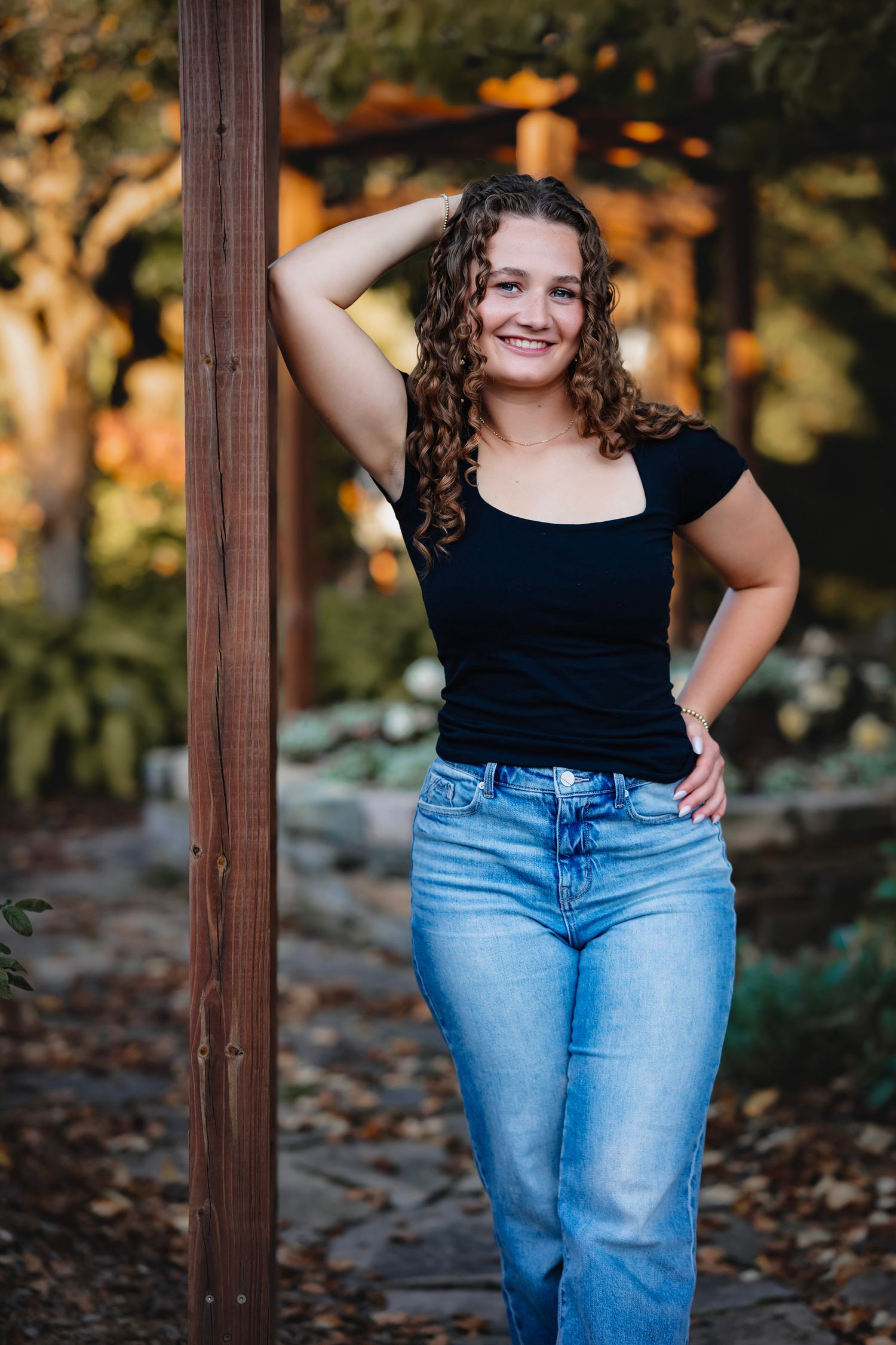 Person in black top and blue jeans poses confidently against wooden post during autumn photoshoot.