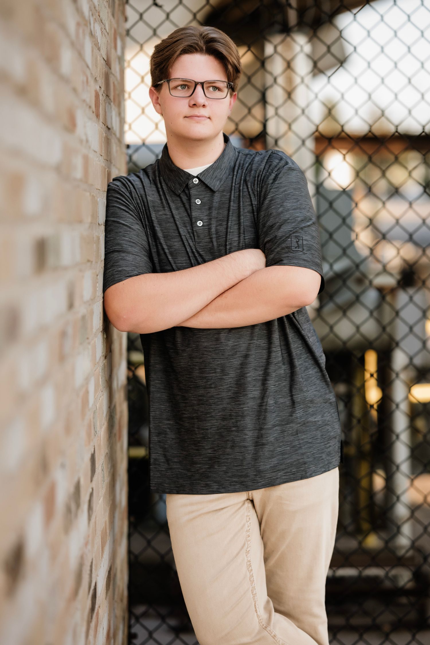 Portrait of a person in glasses wearing a dark gray shirt and khaki pants leaning against a chain link fence.