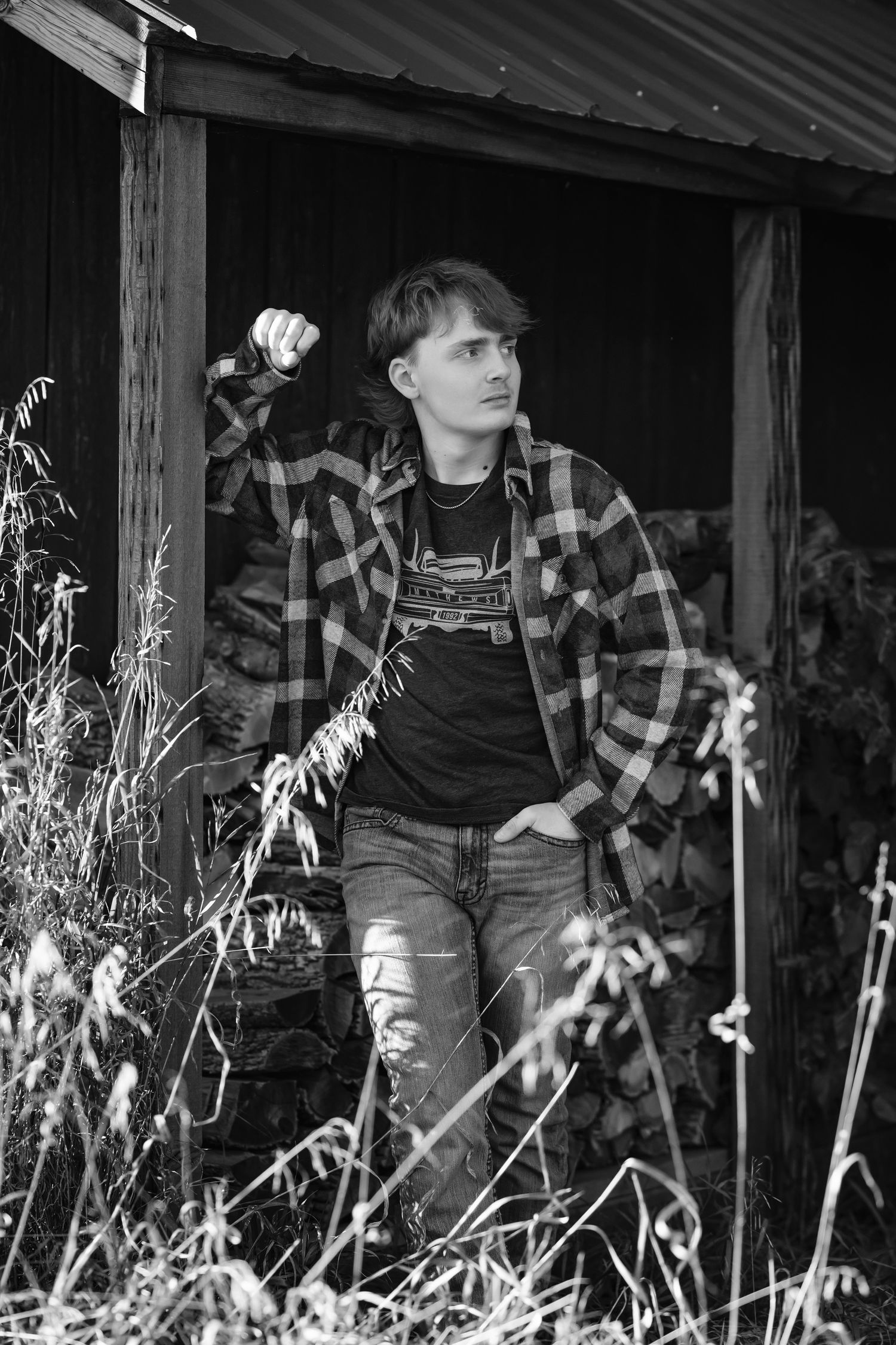 Black and white photo showing someone in plaid flannel and overalls sitting on a rustic wooden porch with dried plants.