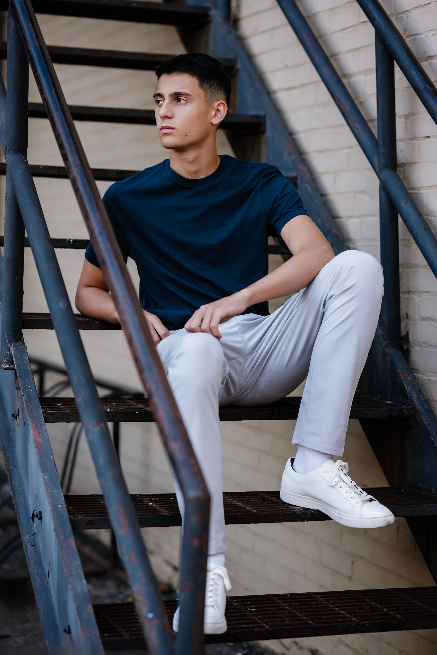 Person in black shirt and white pants sits casually on metal staircase wearing white sneakers in artistic portrait series.