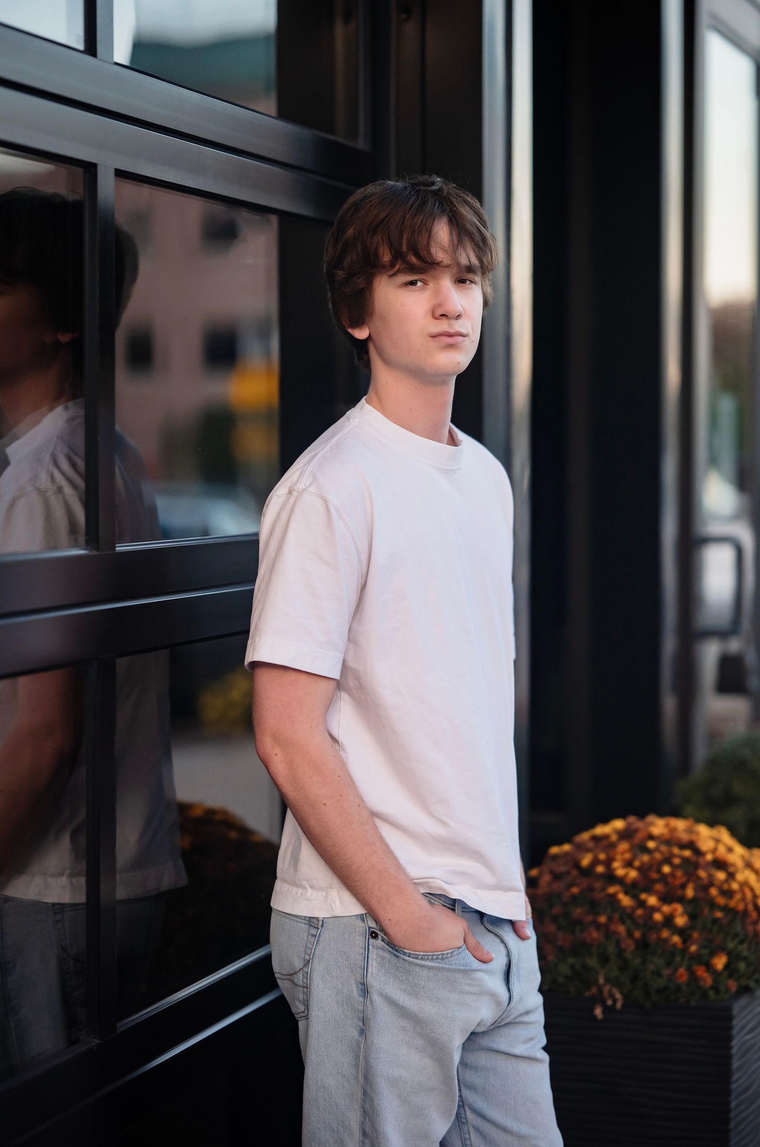 Person in white t-shirt and light jeans stands against dark modern building exterior with orange flowers in planter.