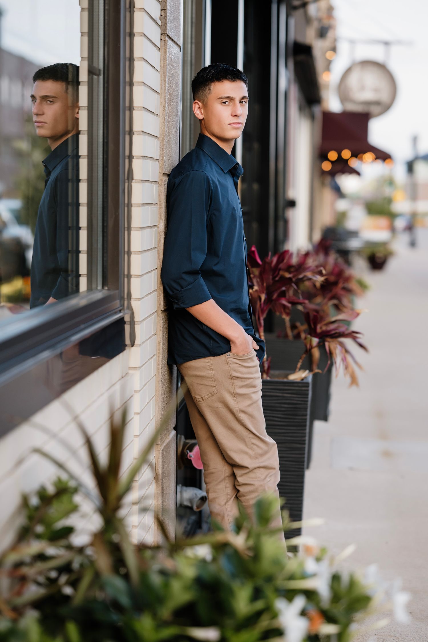 Young man in navy shirt and khaki pants leaning against brick wall on urban sidewalk with plants.