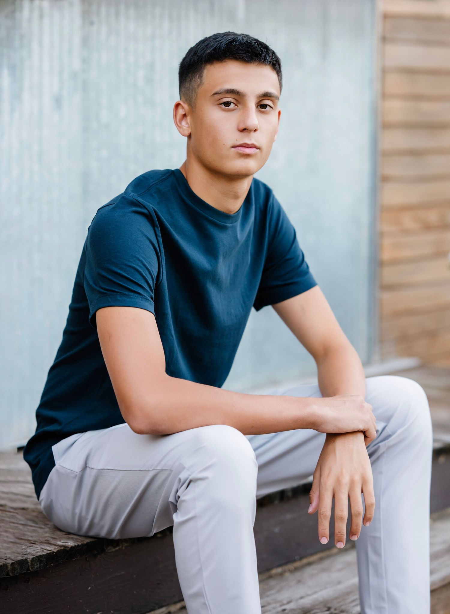 Young man in navy t-shirt and gray pants sitting casually on wooden steps outdoors.