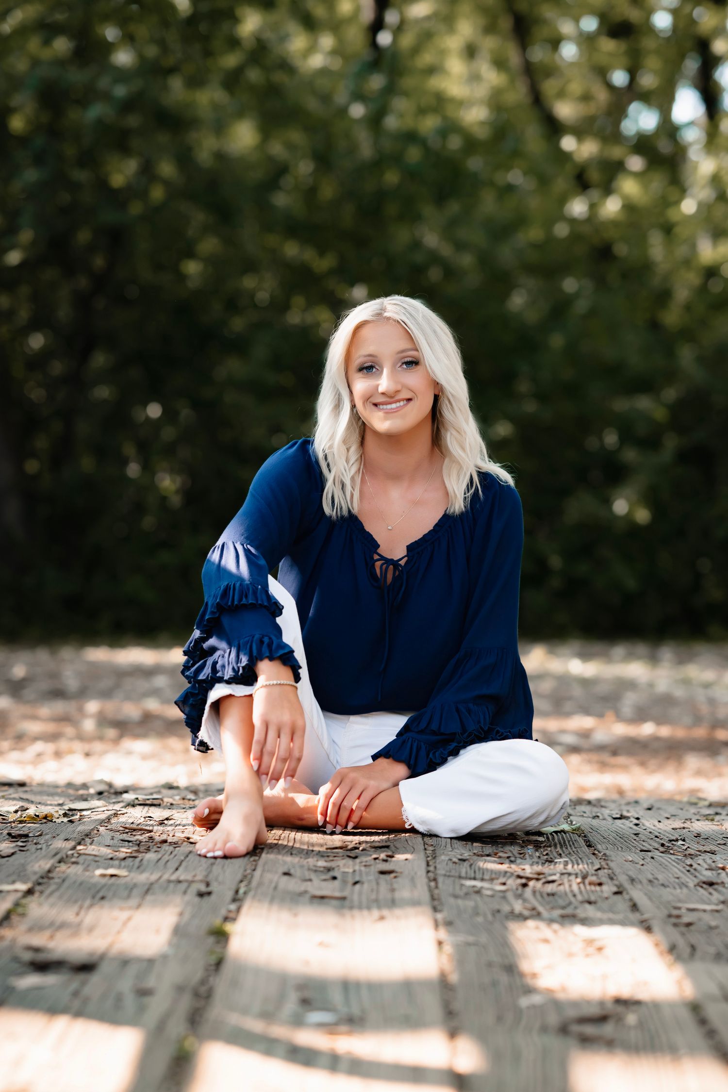 Smiling blonde woman in navy blue top and white pants sitting cross-legged on a wooden deck outdoors.
