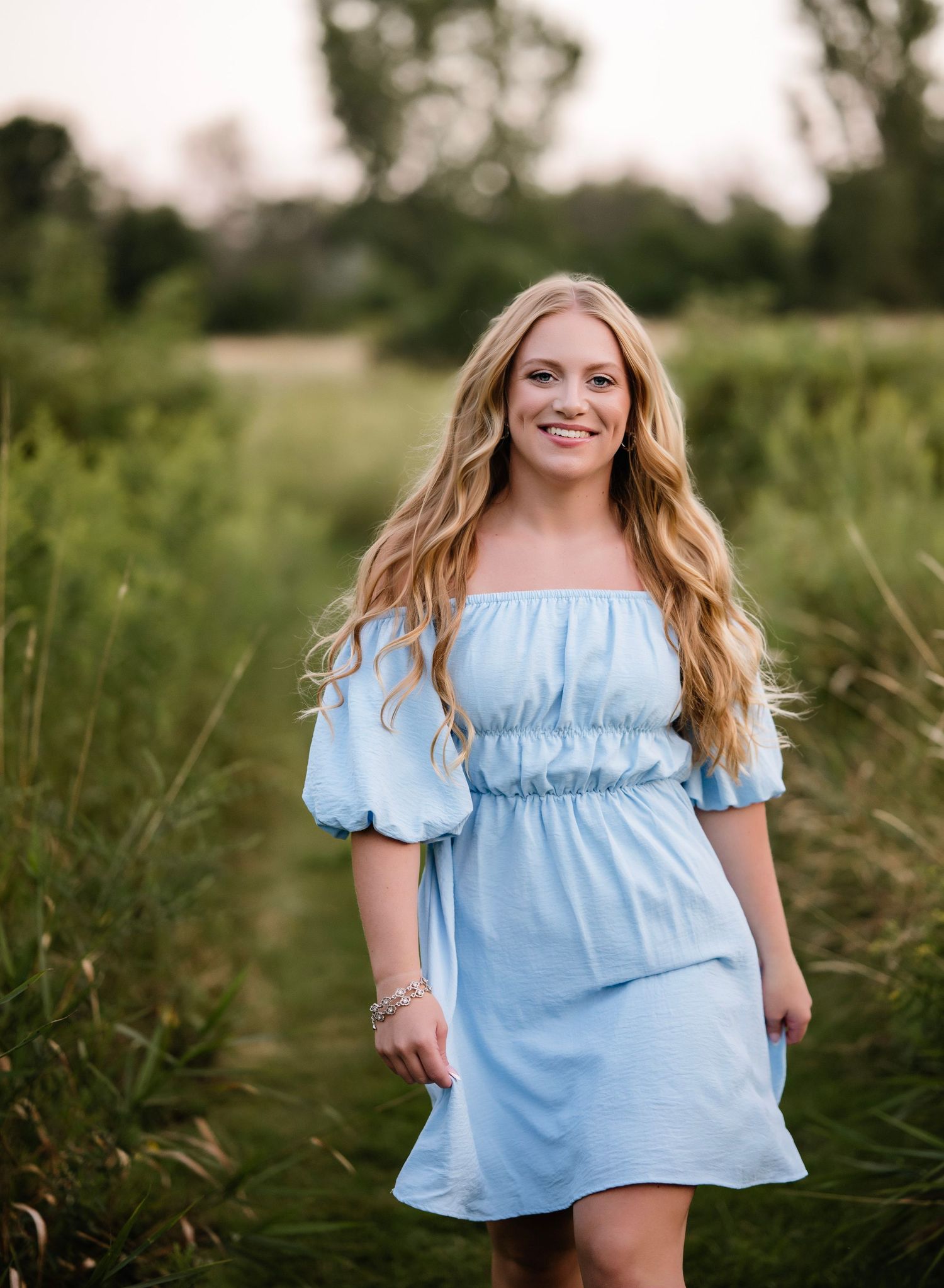 A person in a light blue off-shoulder dress stands in a lush green field during golden hour.