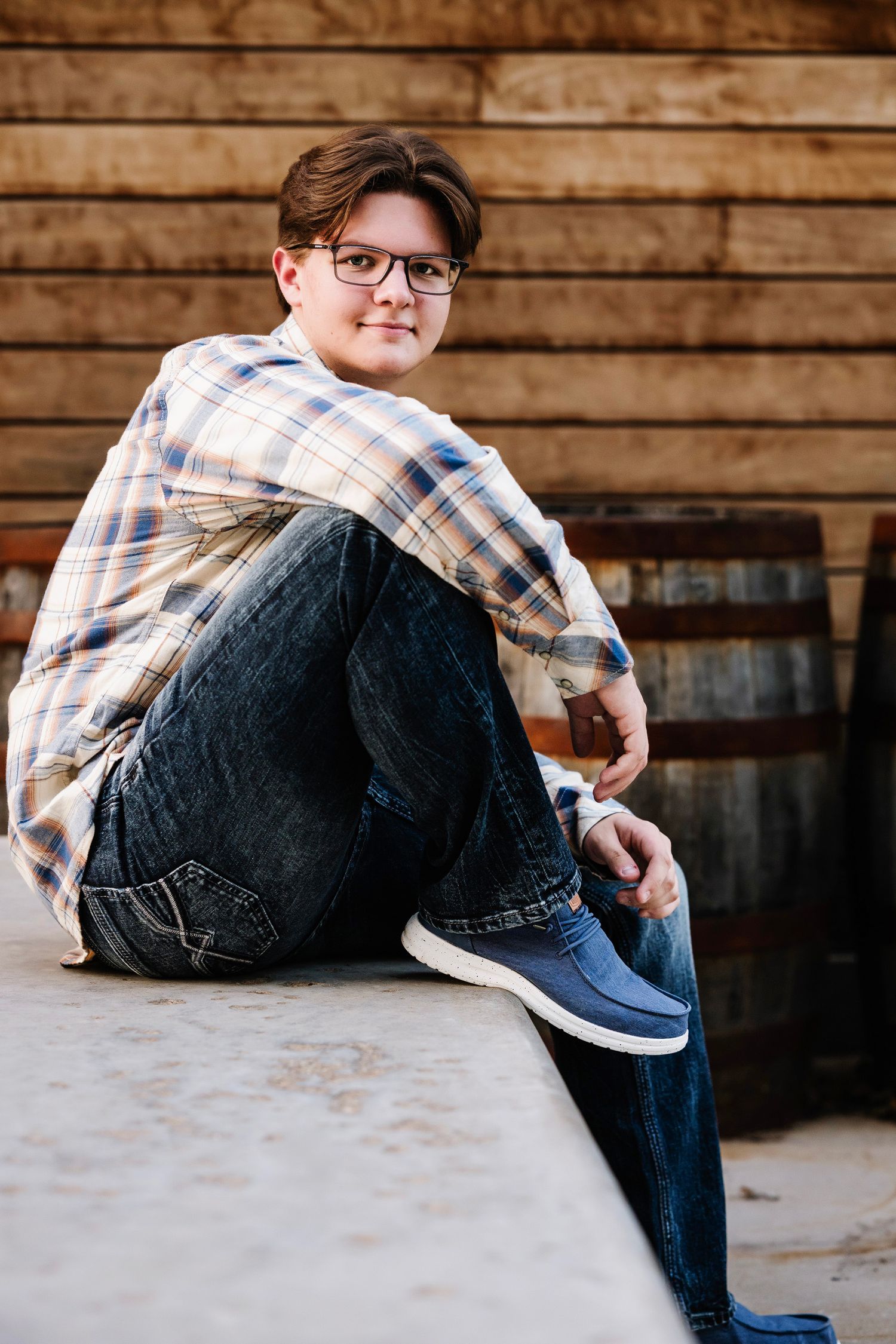 Person in plaid shirt and jeans sitting on concrete surface with wooden wall background in casual pose.