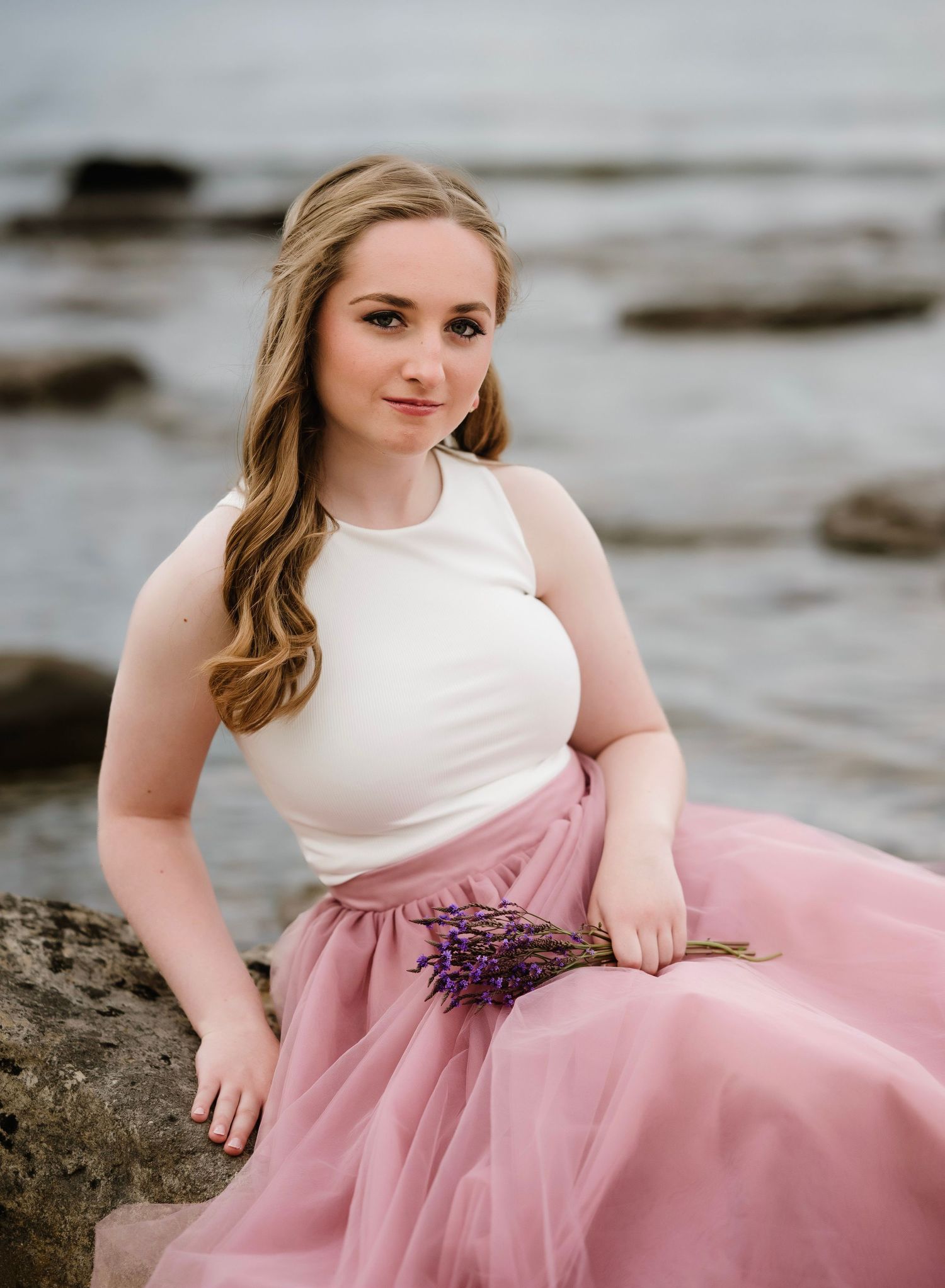 A figure in a white top and flowing pink tulle skirt poses gracefully on rocky coastal terrain at sunset.