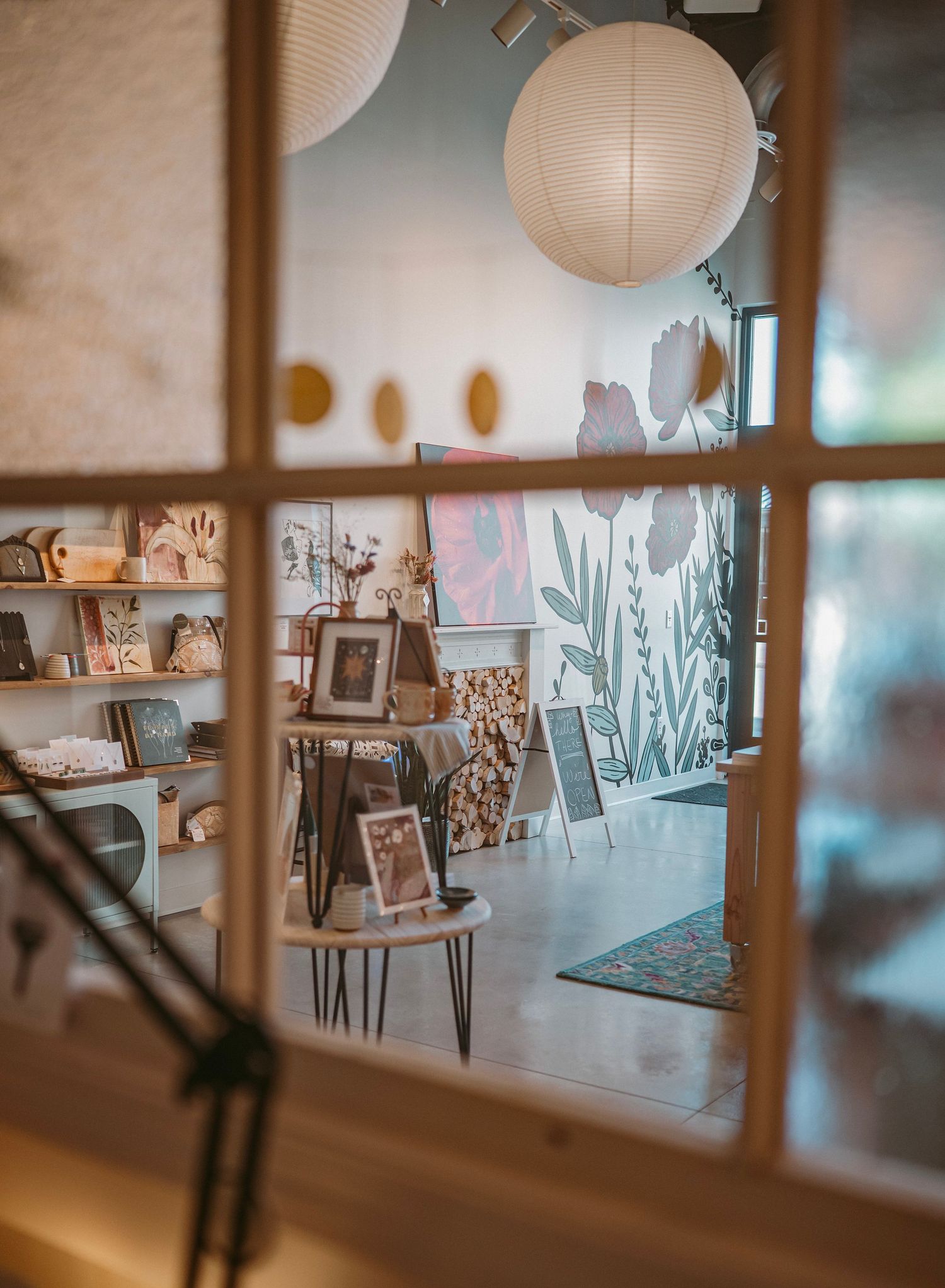 View through window panes into art gallery space with paper lanterns, framed artwork and botanical prints on display.