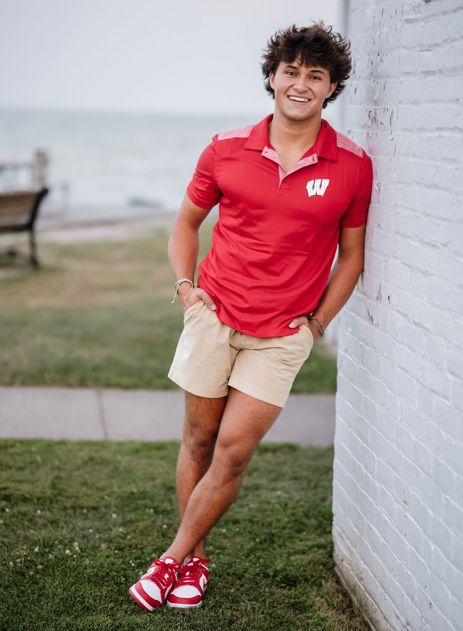 A person in a red Wisconsin polo and khaki shorts leans casually against a white brick wall outdoors.
