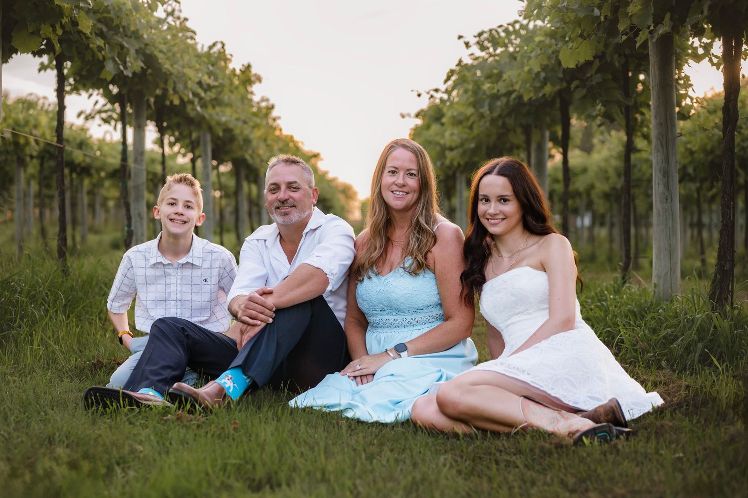 Family of four smiling together in a lush vineyard, dressed in elegant casual attire for a portrait.