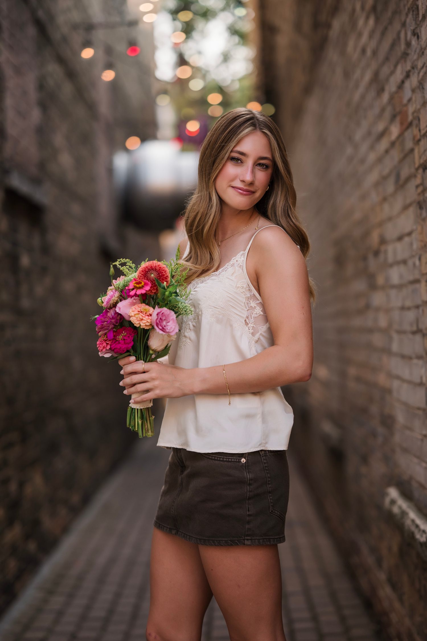 Appleton high school senior holding colorful pink bouquet in white cami and dark shorts, posing in a brick alley with bokeh lights.