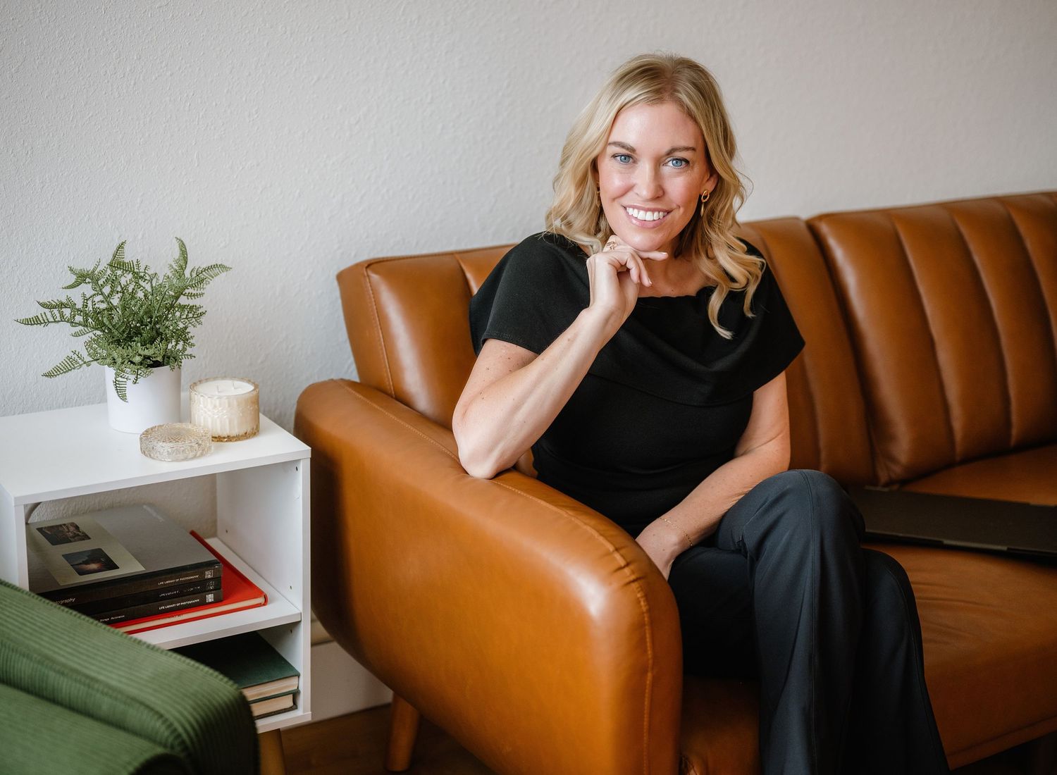 A person sits casually on a tan leather couch while wearing all black clothing in a bright minimalist living space.