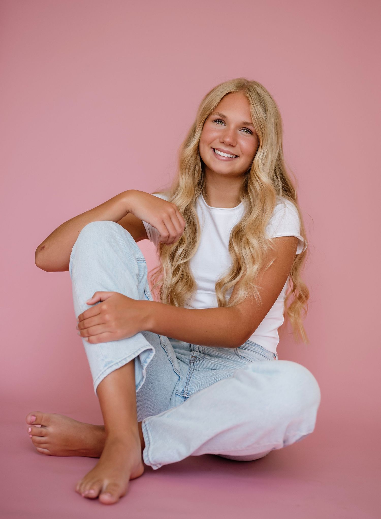 Smiling blonde woman in white tee and light jeans sitting barefoot against a pink background.