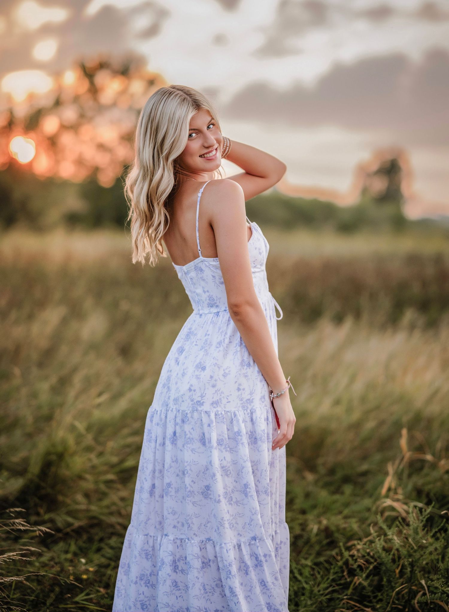 Blonde woman in white floral maxi dress posing in golden hour sunlight in a lush green field.
