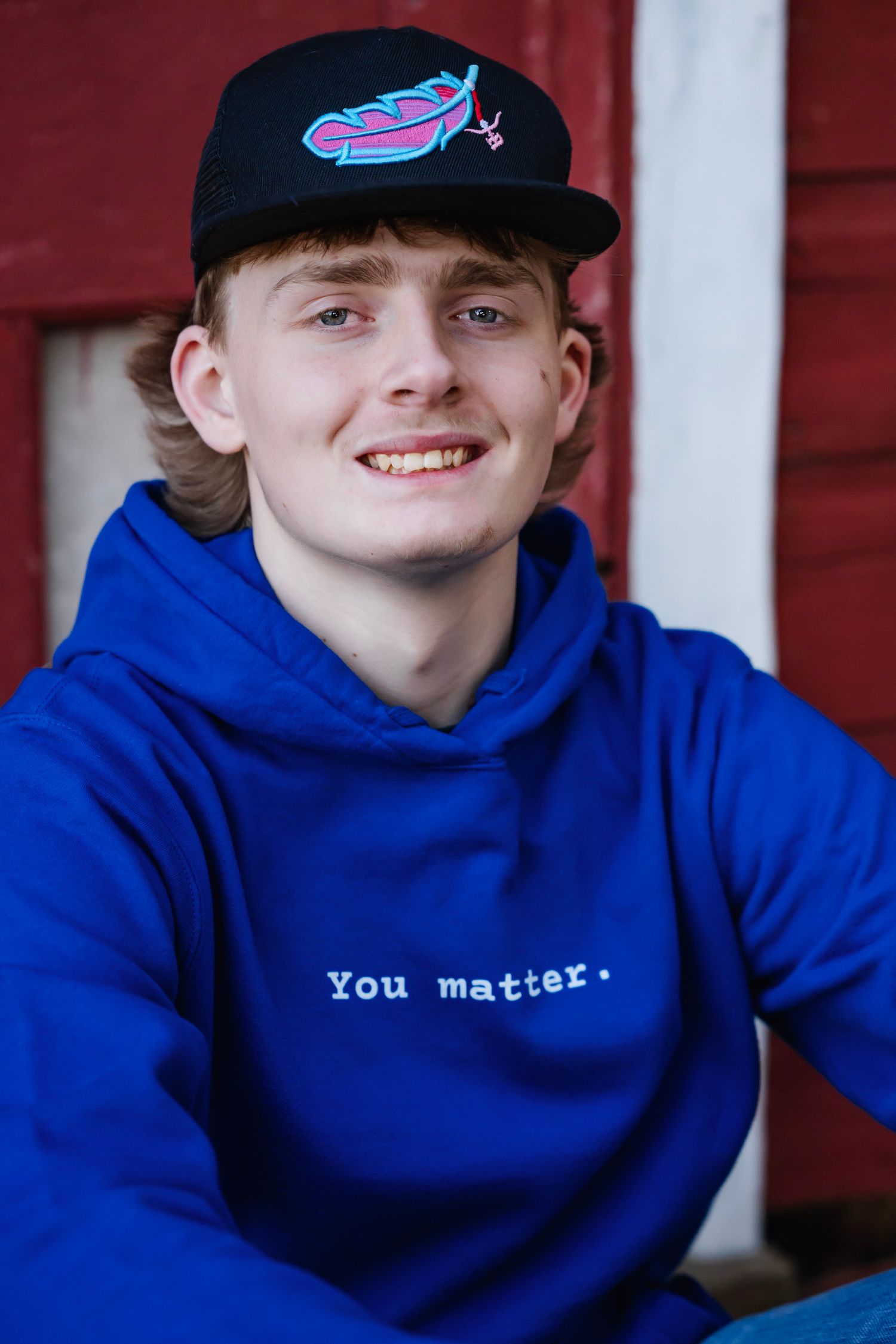 A person in a blue hoodie and baseball cap smiles while sitting against a red wall background.
