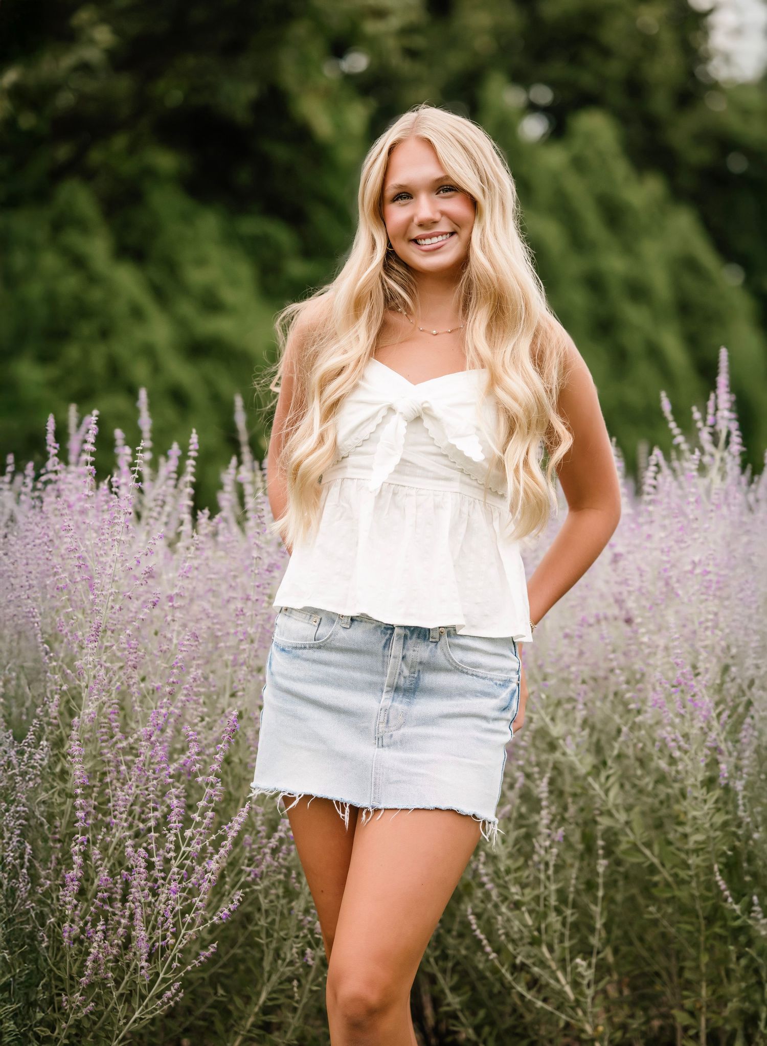 A person in a white top and denim shorts poses in a field of blooming purple Russian sage flowers on a summer day.