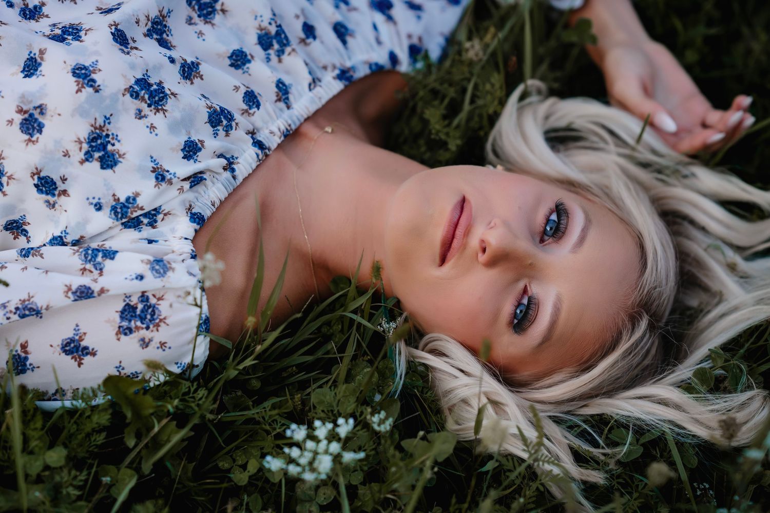 Person with blonde hair lying in grass wearing a floral print dress with blue flowers against a dark background.