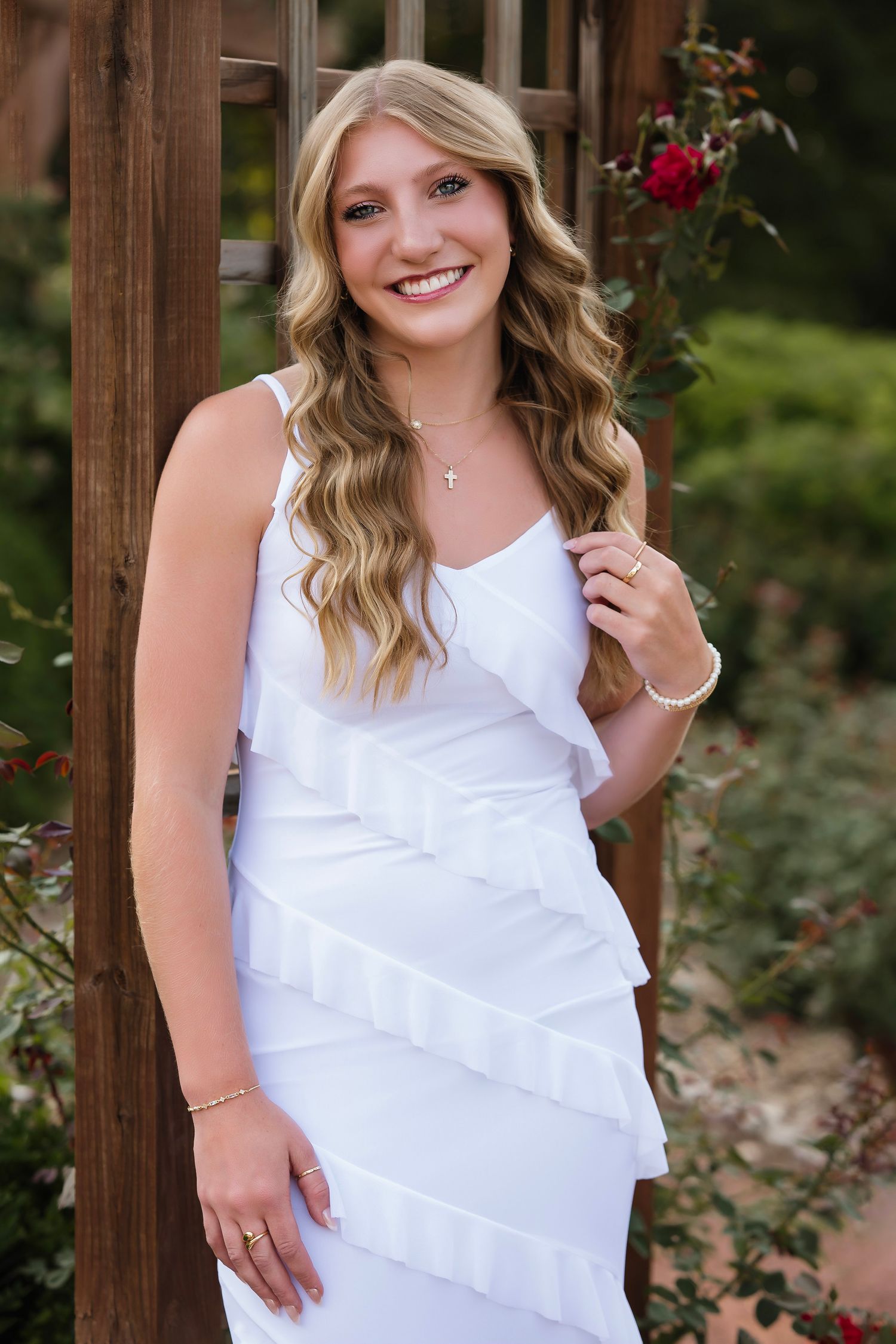 Smiling young woman in white ruffled dress posing by wooden arbor with red roses in garden background.