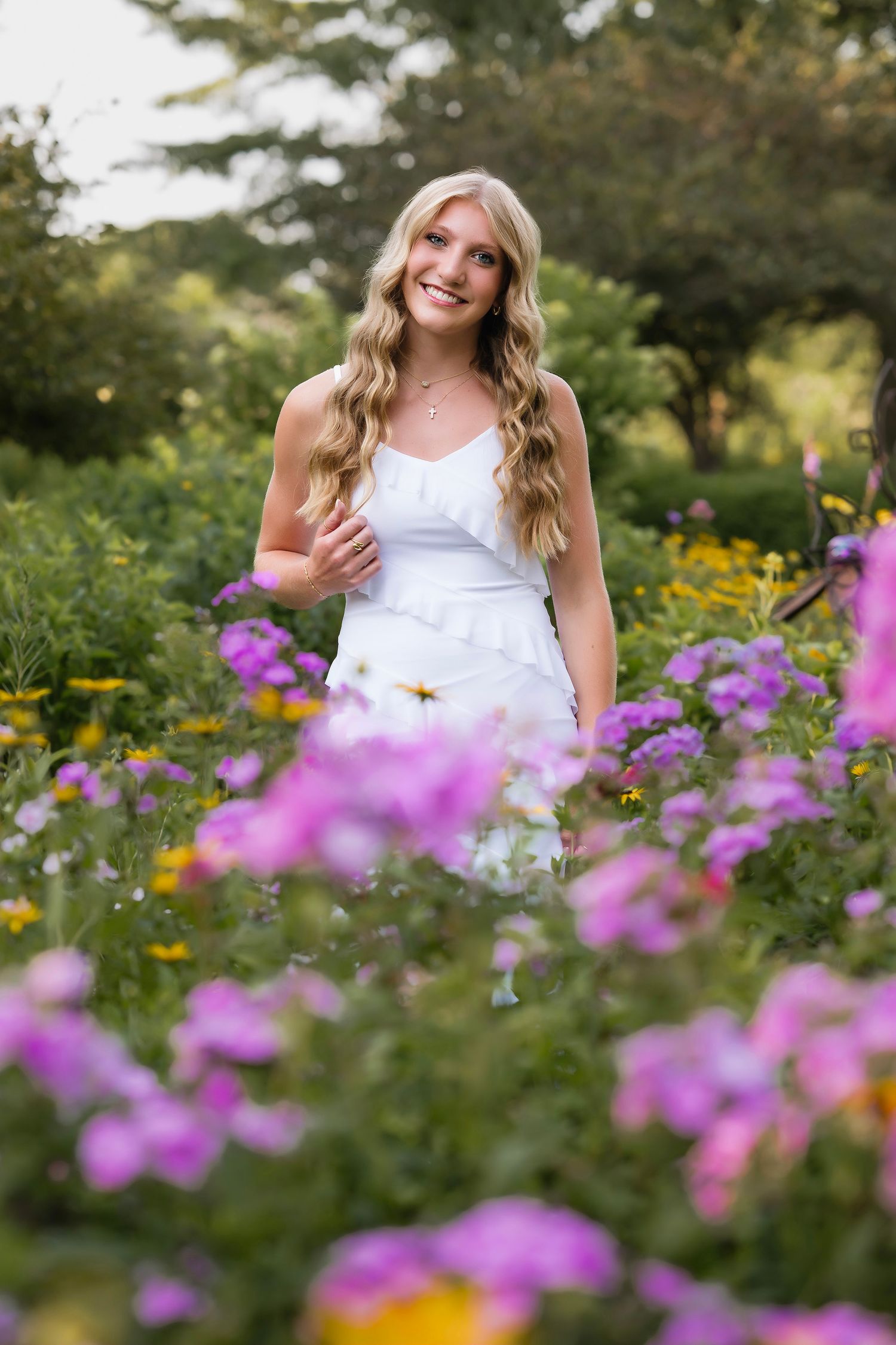 Smiling blonde woman in white dress standing among vibrant purple and yellow wildflowers in a lush garden.