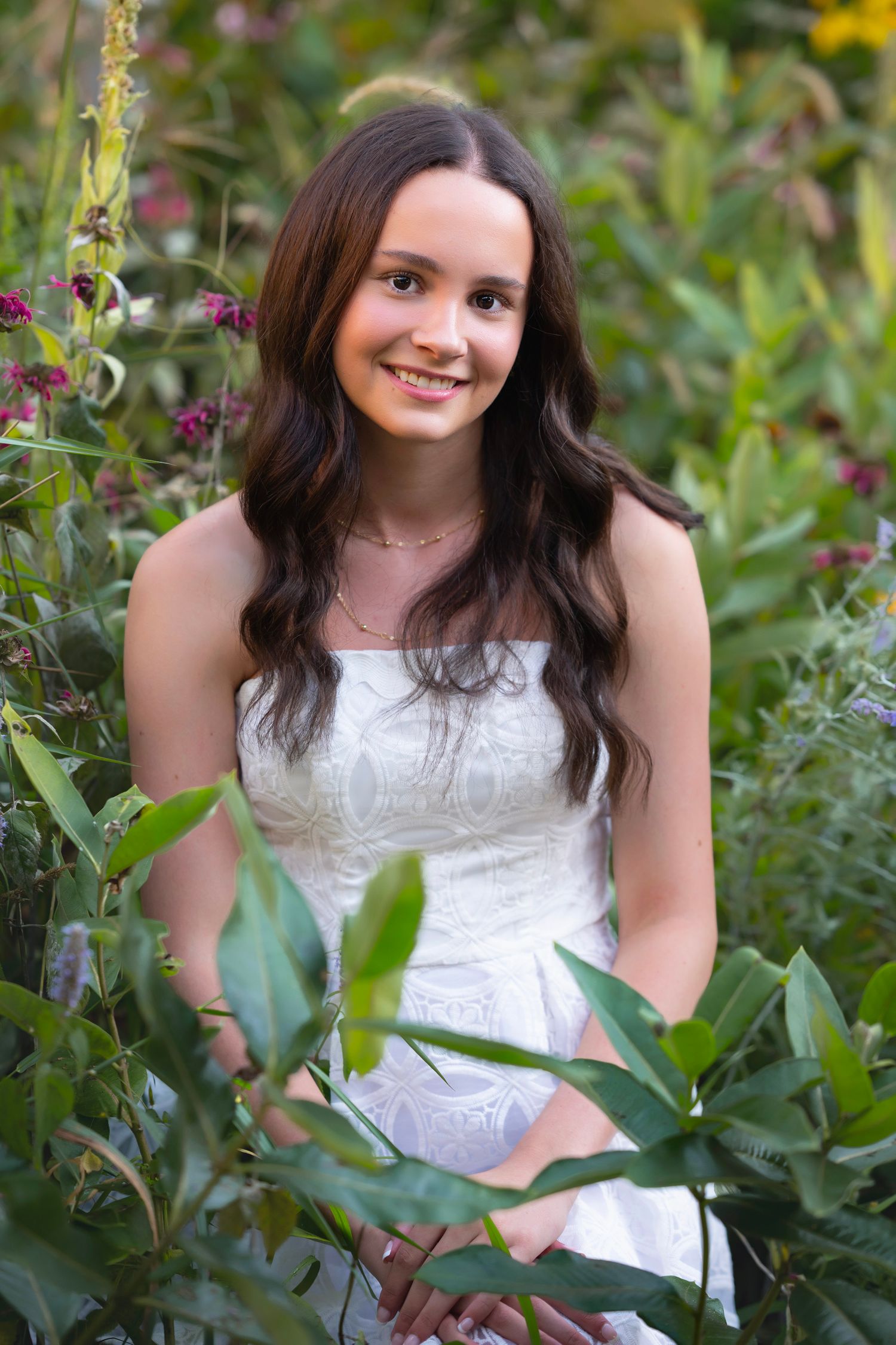 Young woman in white strapless dress smiling among lush green garden plants and pink wildflowers.