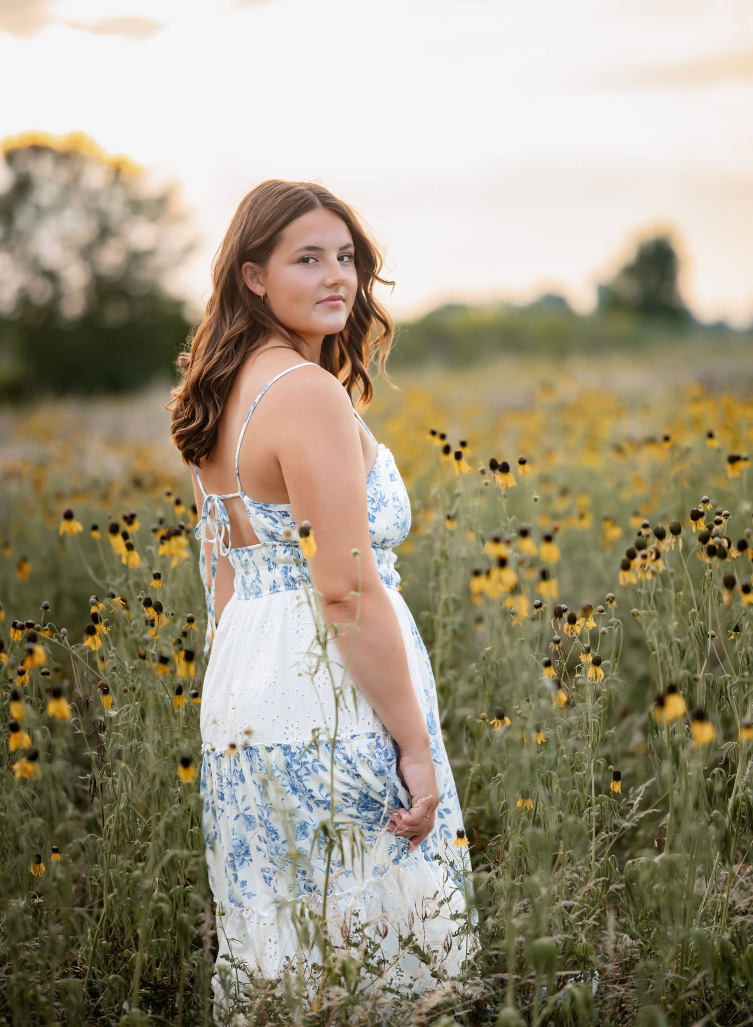 A person in a floral dress sits gracefully among blooming yellow wildflowers in a field at sunset.