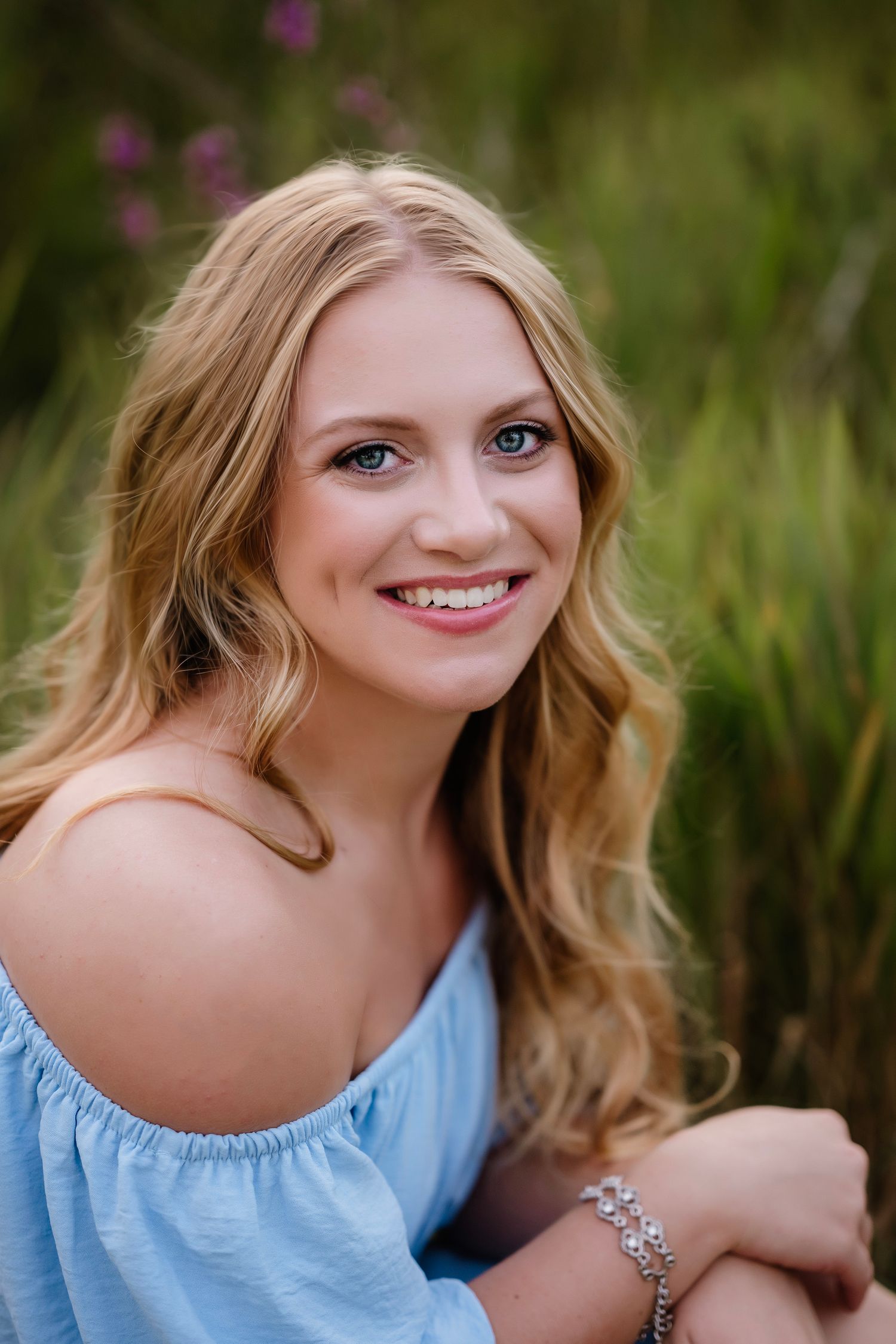 A person in a light blue off-shoulder top poses outdoors against a natural green background in summer.