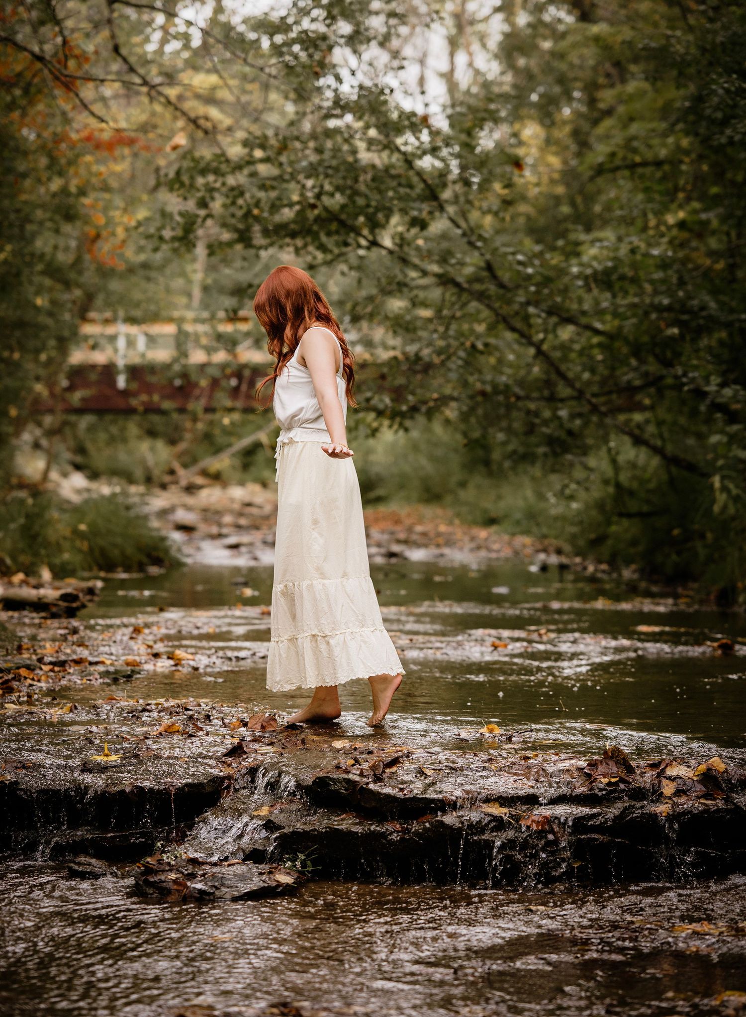 A figure in a flowing white dress stands gracefully on rocks by a tranquil stream in a wooded setting during autumn.