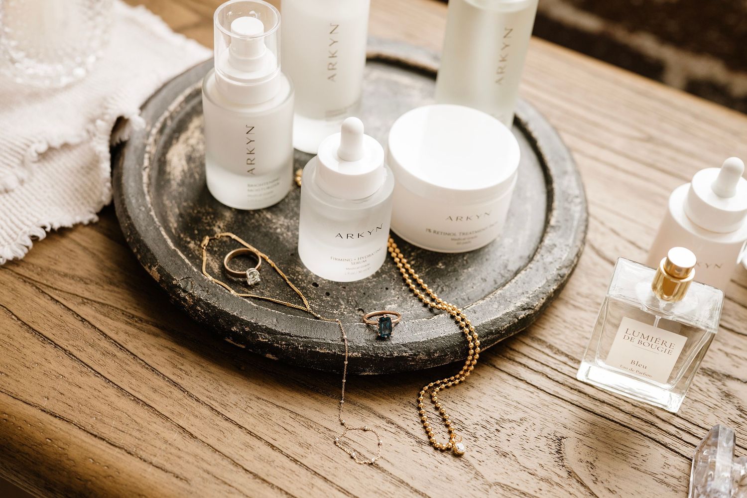 White skincare products and gold jewelry arranged on a rustic wooden tray beside a perfume bottle.
