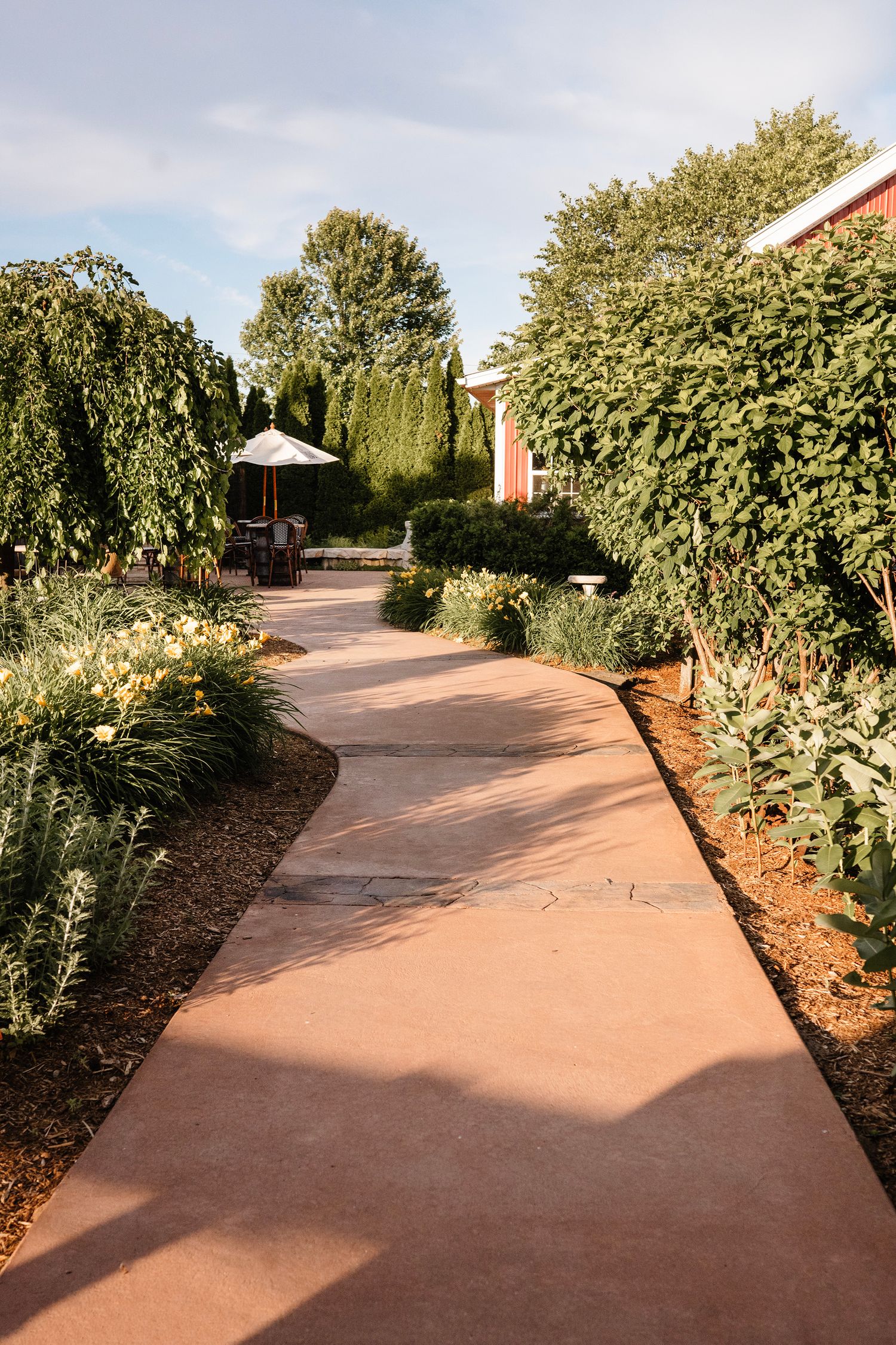 A winding red brick pathway through a lush garden with manicured shrubs, flowering plants, and a patio with umbrella.