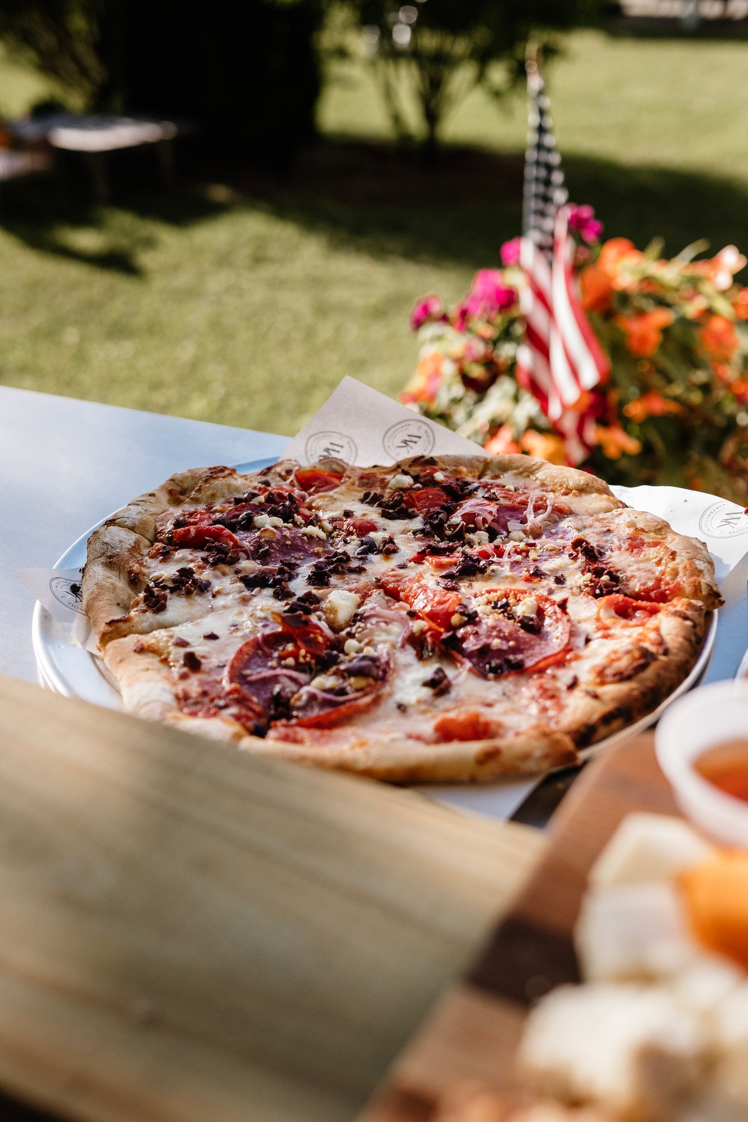 Outdoor pizza on a metal pan with tomatoes, cheese, and toppings, with an American flag and flowers in the background.