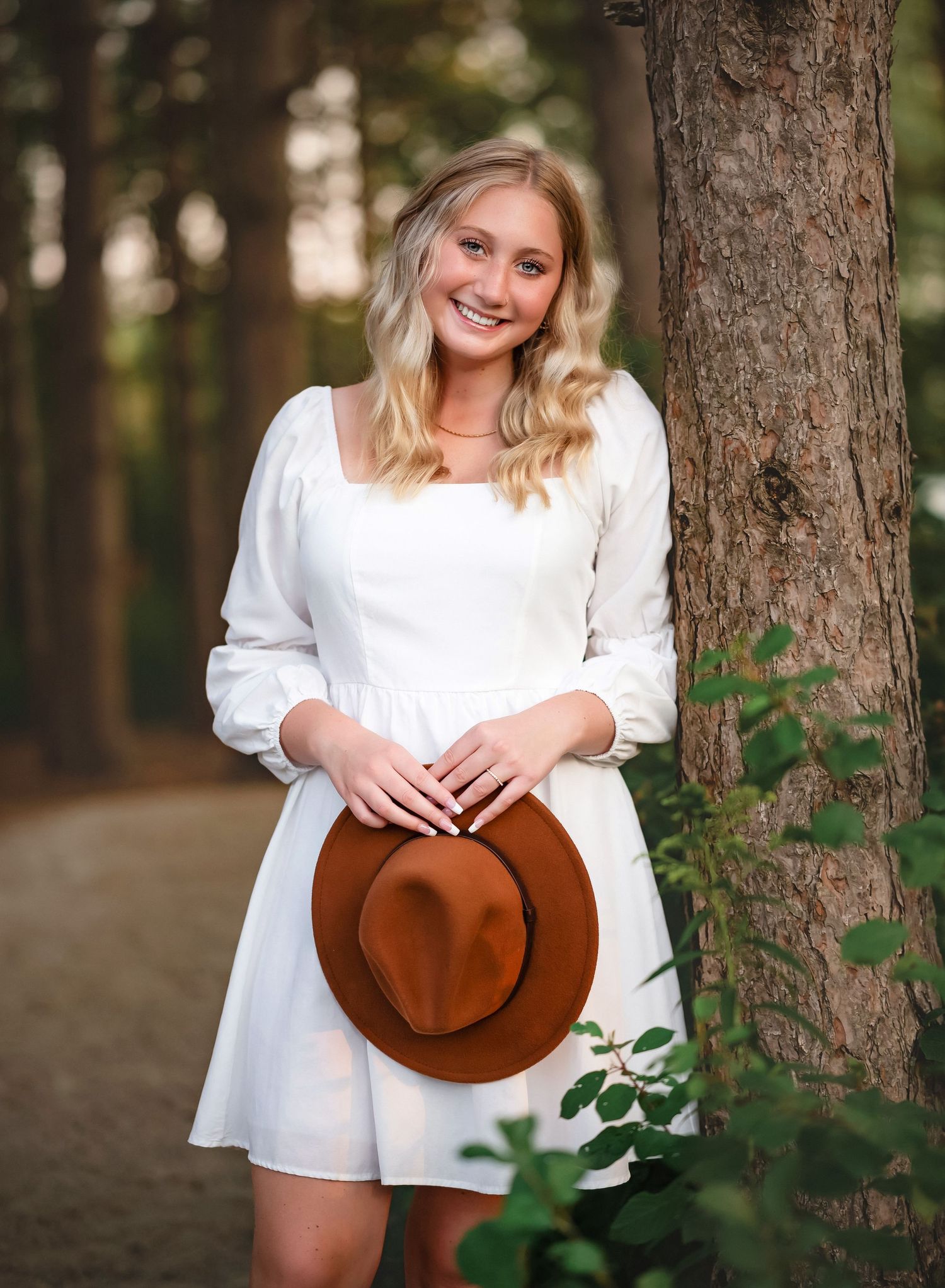 A white summer dress and brown felt hat are showcased in an outdoor forest high school senior portrait against tree trunks and greenery.