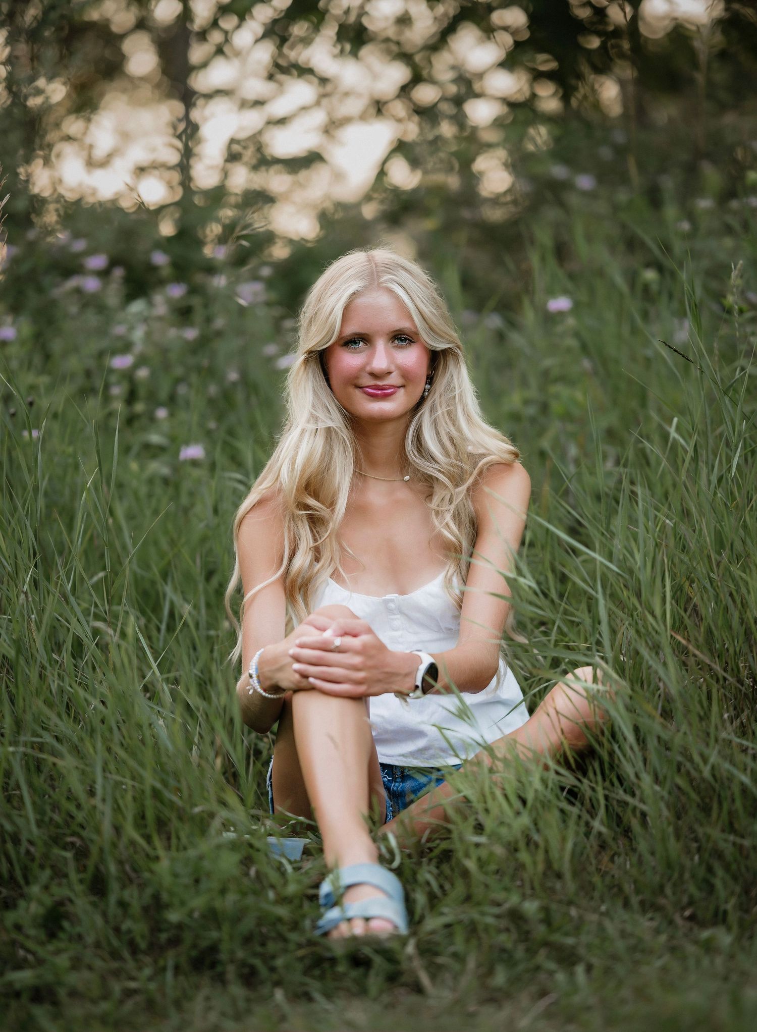 A person sits in tall grass at sunset wearing a white top and denim shorts with soft bokeh lighting in the background.