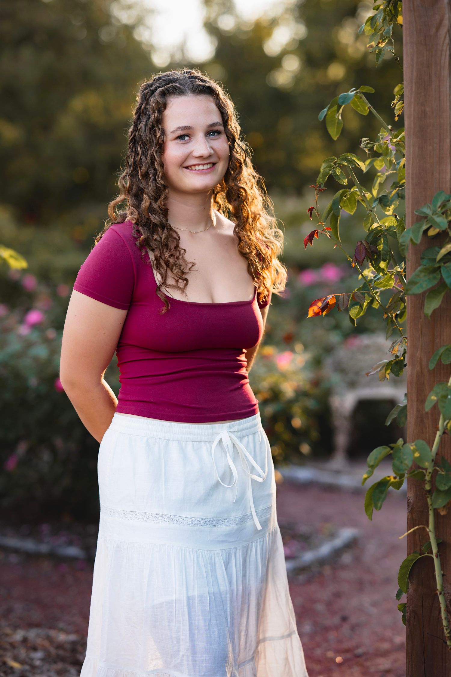 A person in a burgundy top and white skirt poses outdoors in a garden setting with flowers and greenery.