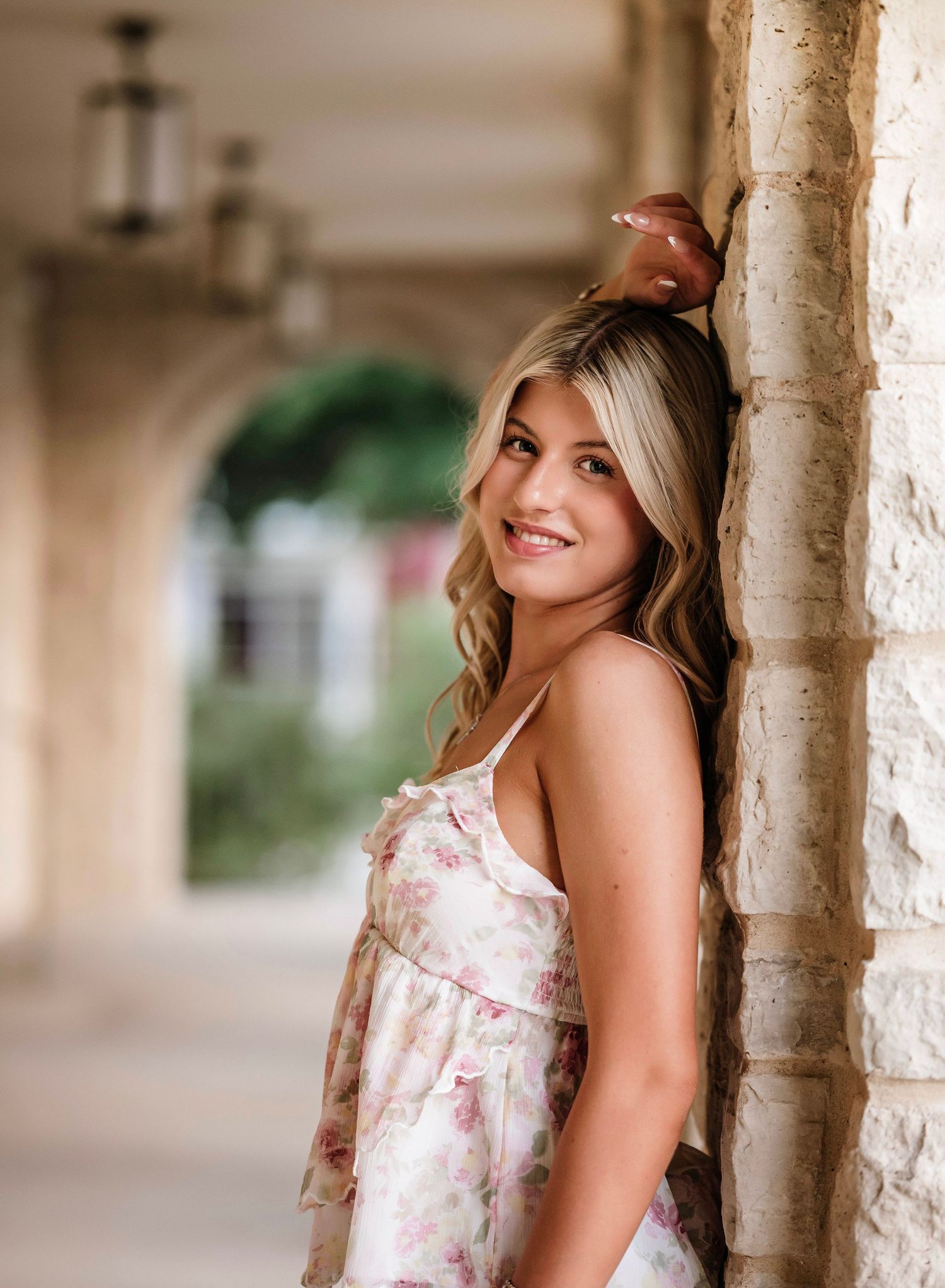 A woman in a floral dress poses against a stone pillar in a covered walkway with soft natural lighting.