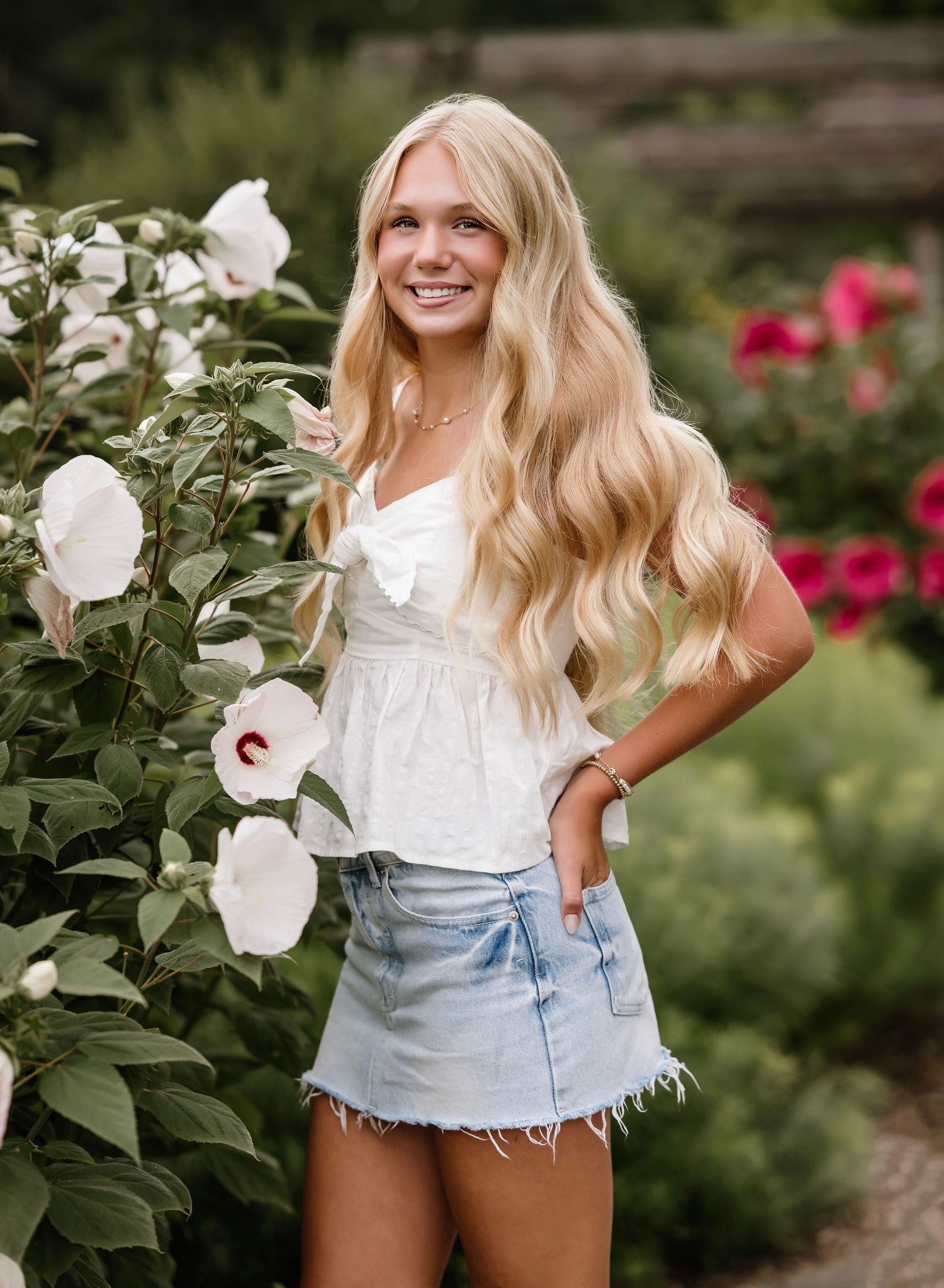 Smiling blonde teen in white top and denim skirt posing beside white hibiscus flowers in a lush garden.