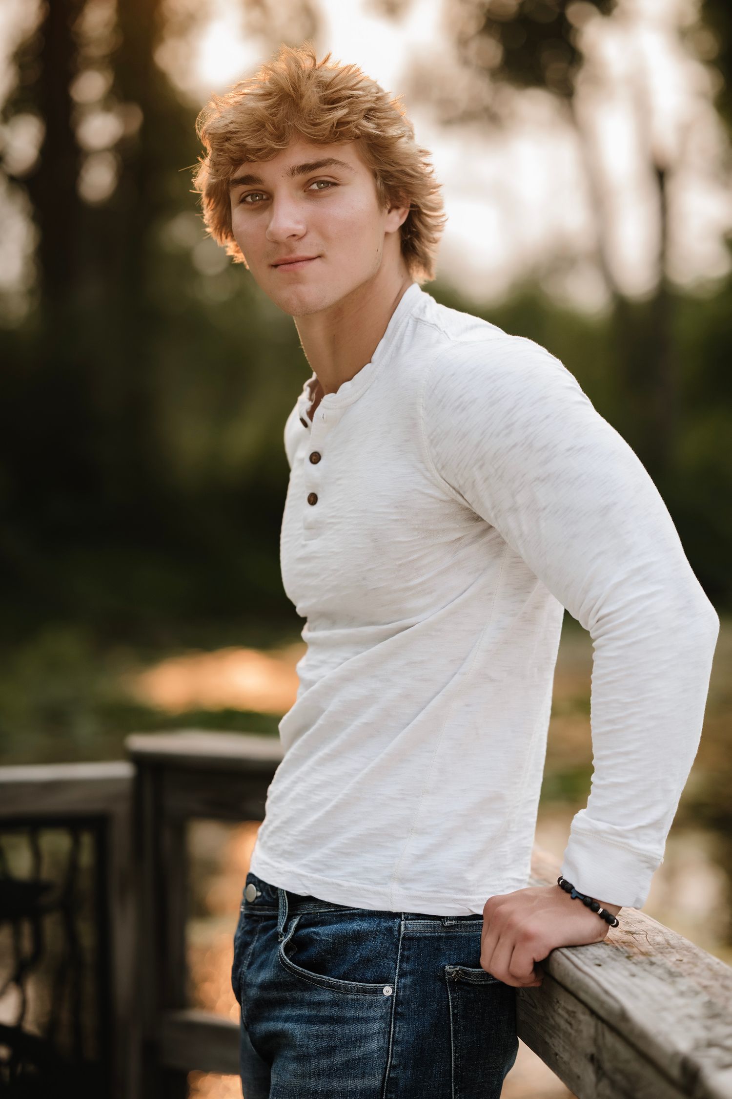 Young man with blond hair in white henley shirt and jeans leaning on wooden railing outdoors.