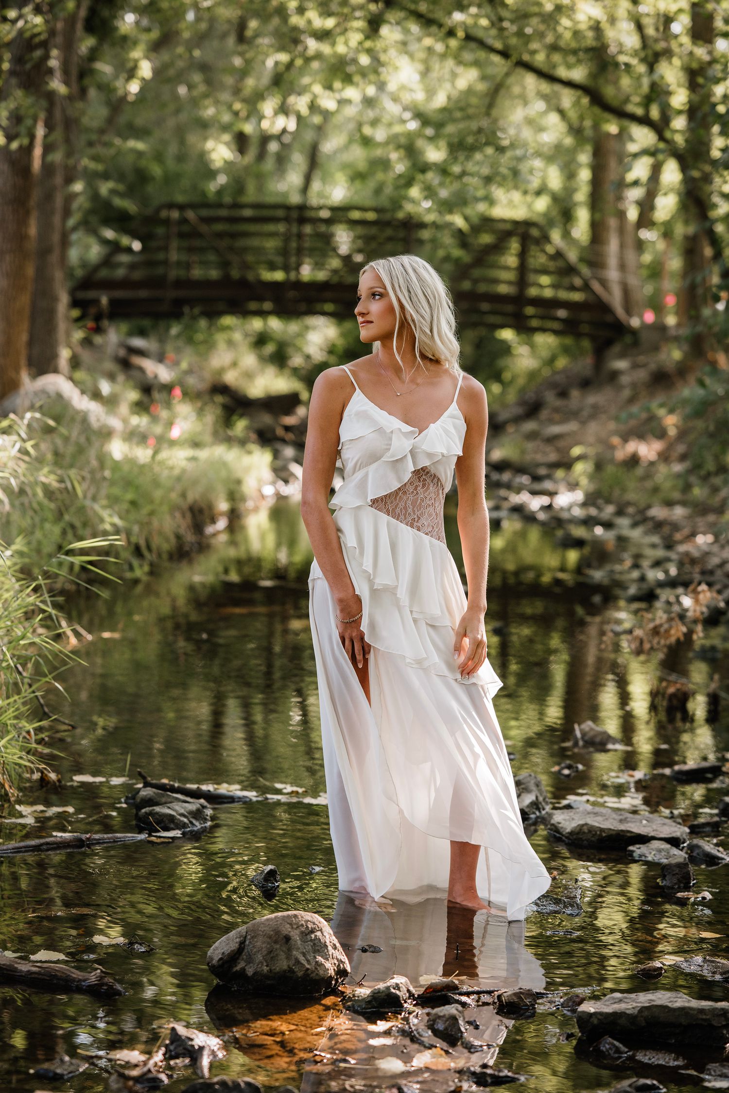 A ethereal figure in a white dress stands gracefully in a tranquil forest stream with sunlit reflections on the water.