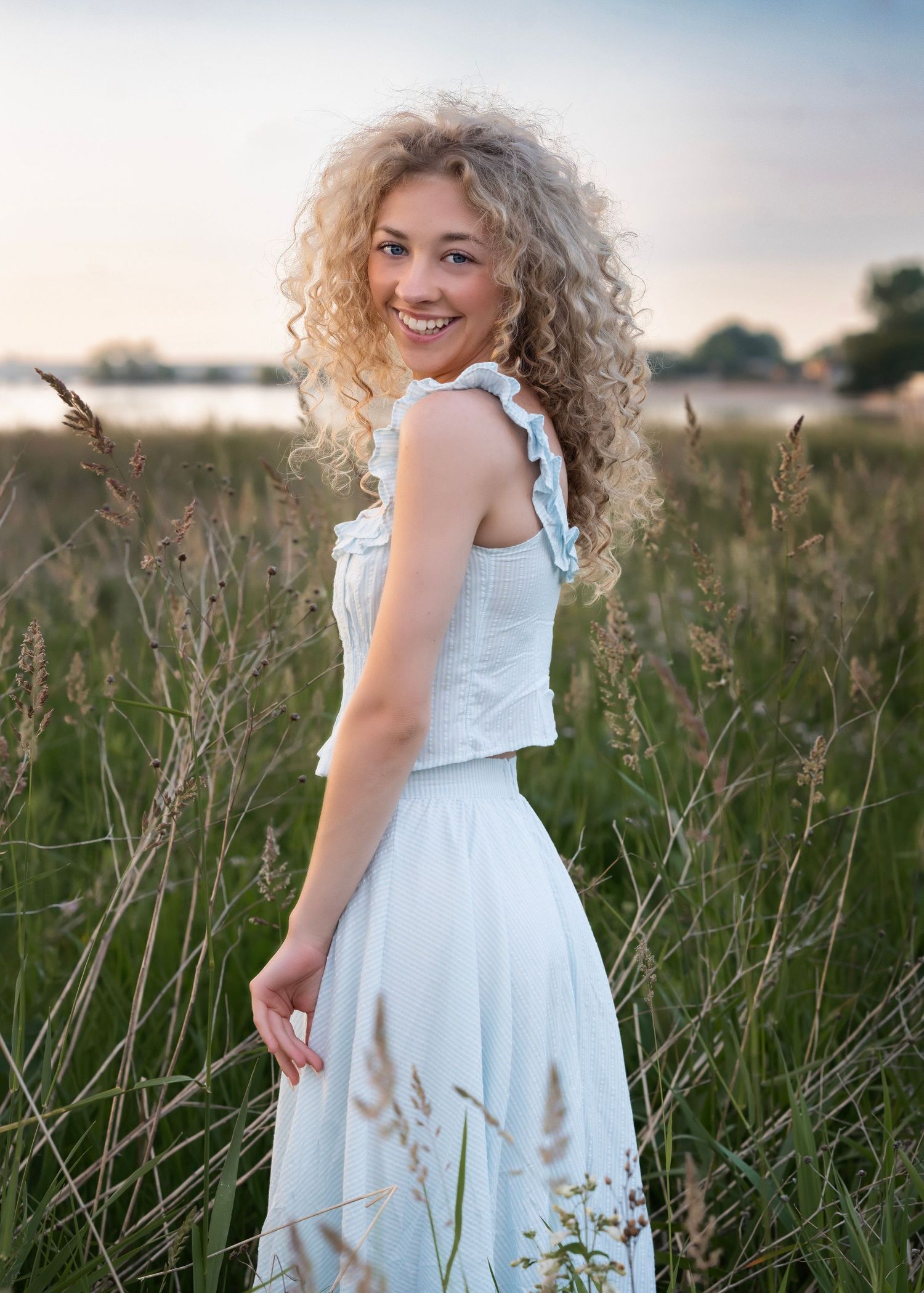 Young woman with curly blonde hair in white dress standing in tall grass field at sunset.