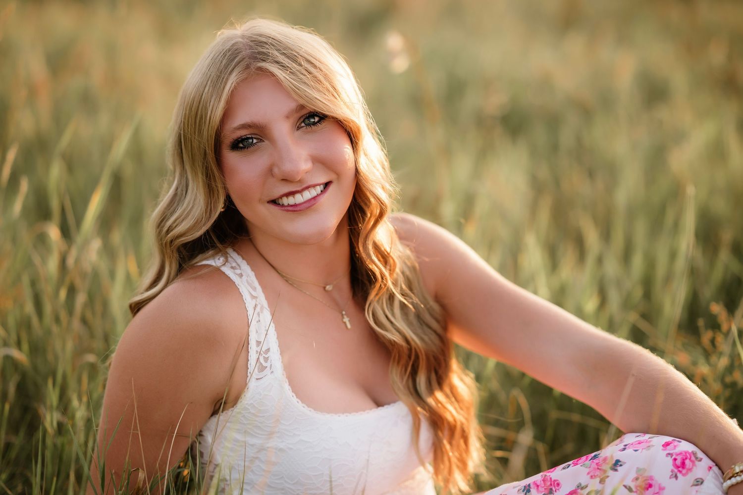 Young woman with long blonde hair smiles warmly while sitting in a golden meadow, wearing a white lace top.