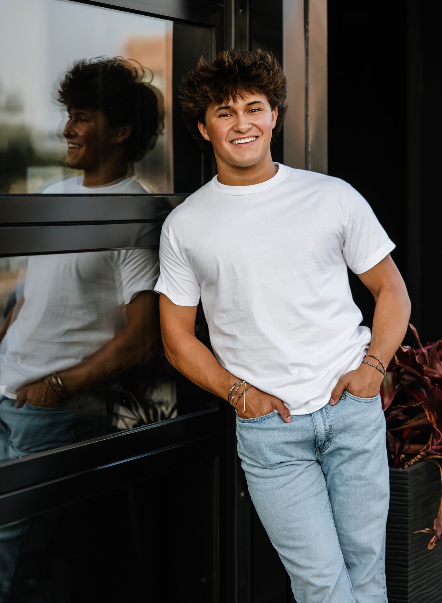 Young person in a white t-shirt and light jeans poses casually against a window with their reflection visible.