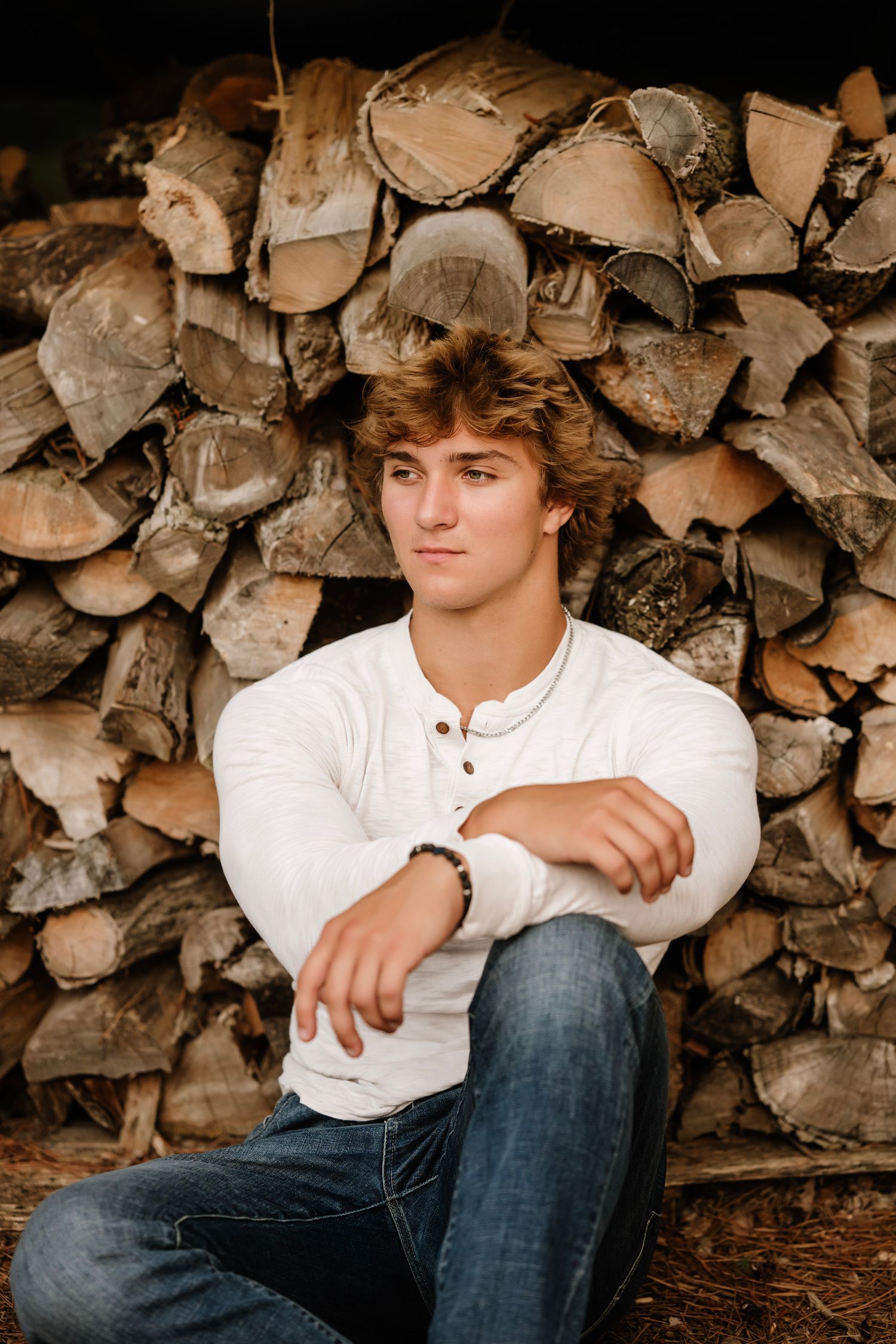 Casual outdoor portrait against rustic woodpile background wearing white henley shirt and jeans.